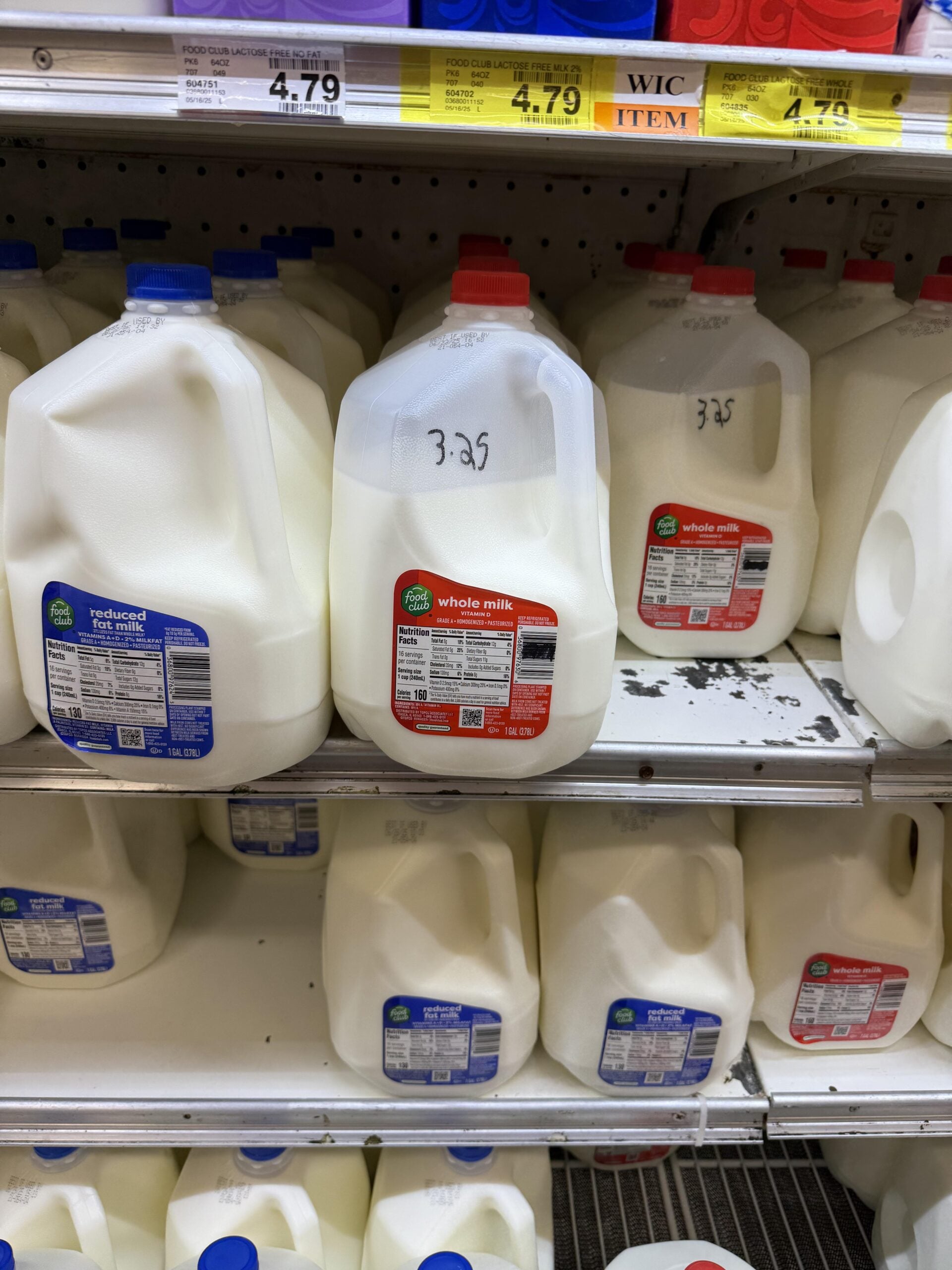 Gallon jugs of whole and reduced fat milk on a grocery store shelf, with price tags reading $4.79 above. Some jugs are turned to show nutrition labels.