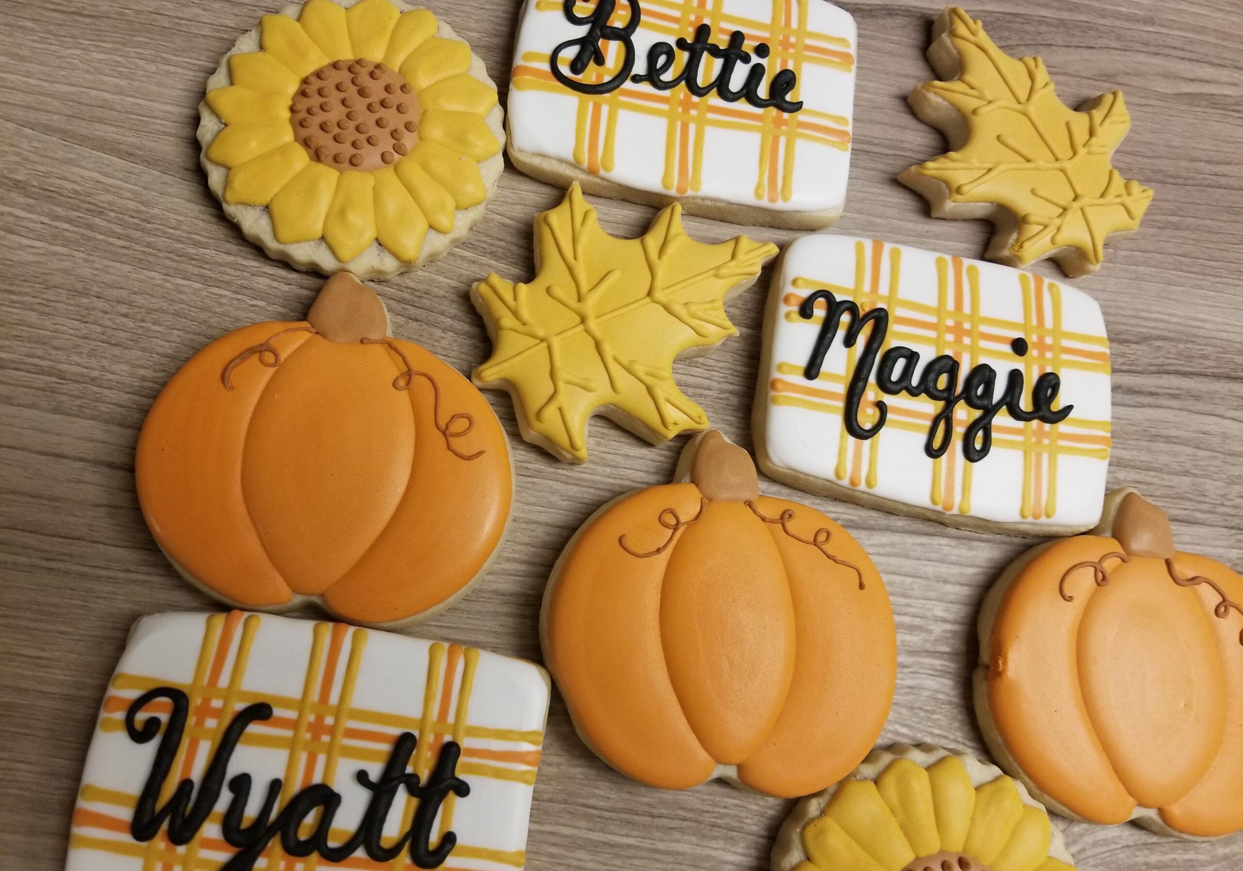 Assorted decorated cookies shaped like pumpkins, sunflowers, and leaves, along with rectangular cookies featuring plaid designs and the names Wyatt, Bettie, and Maggie written in black icing.