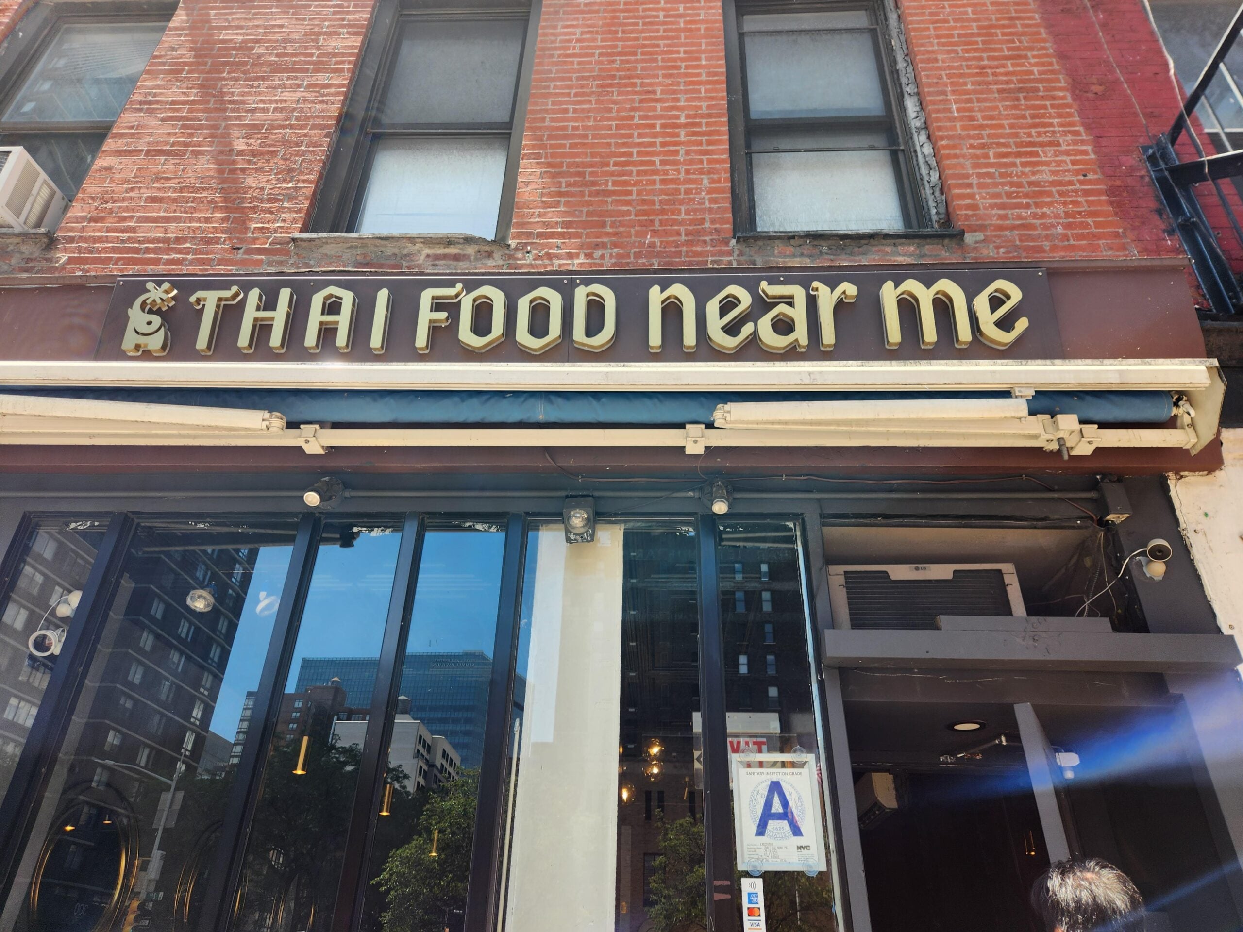 A brick building with a sign reading “THAI FOOD near me” above glass doors. Reflections of nearby buildings are visible in the windows. A health inspection grade “A” sign is displayed by the entrance.