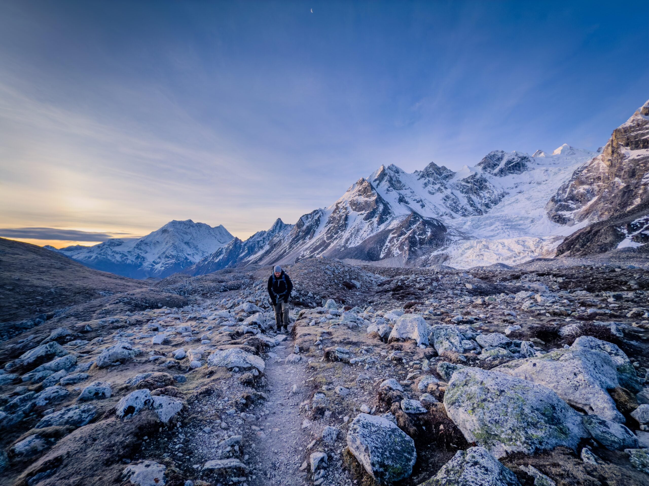 A lone hiker walks along a rocky trail through a barren valley with snow-capped mountains in the background under a clear, blue sky at sunset.