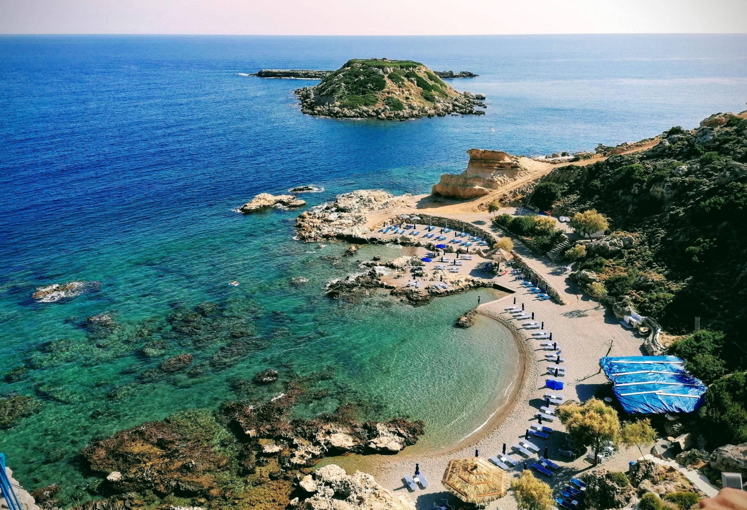 Aerial view of incredible beaches with clear blue water, sun loungers, umbrellas, rocky shoreline, and a small island offshore under a bright sky.