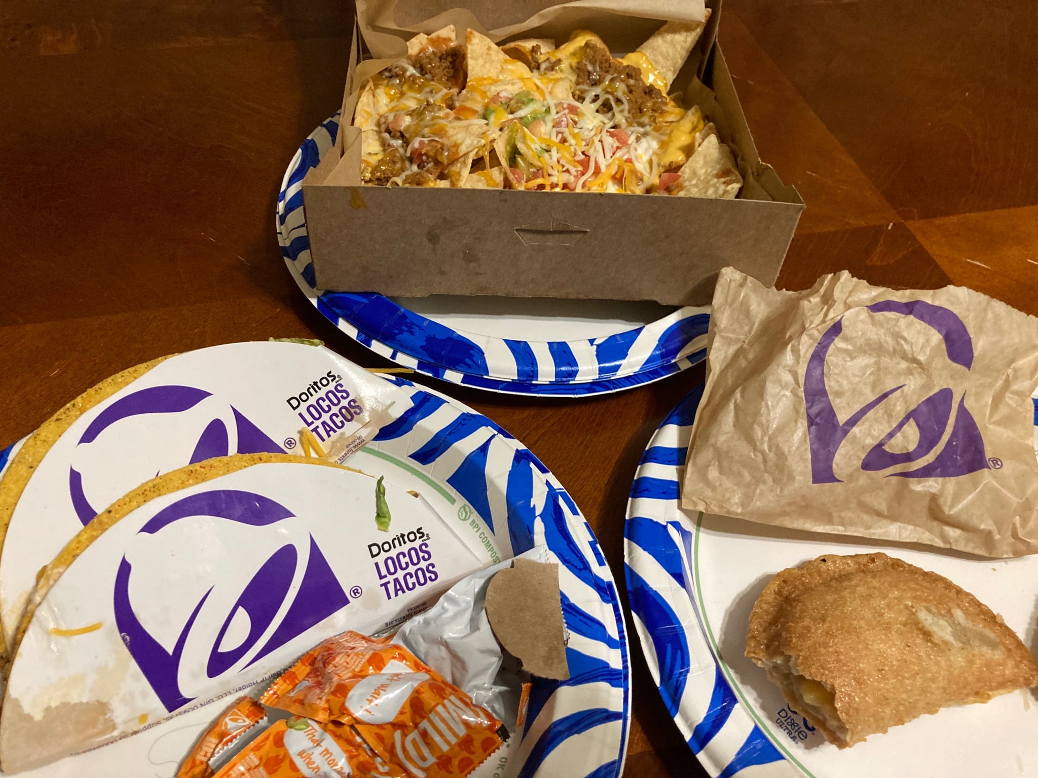 A Taco Bell meal consisting of a box of nachos, two Doritos Locos tacos, a paper bag, and a pastry on blue-and-white patterned plates.