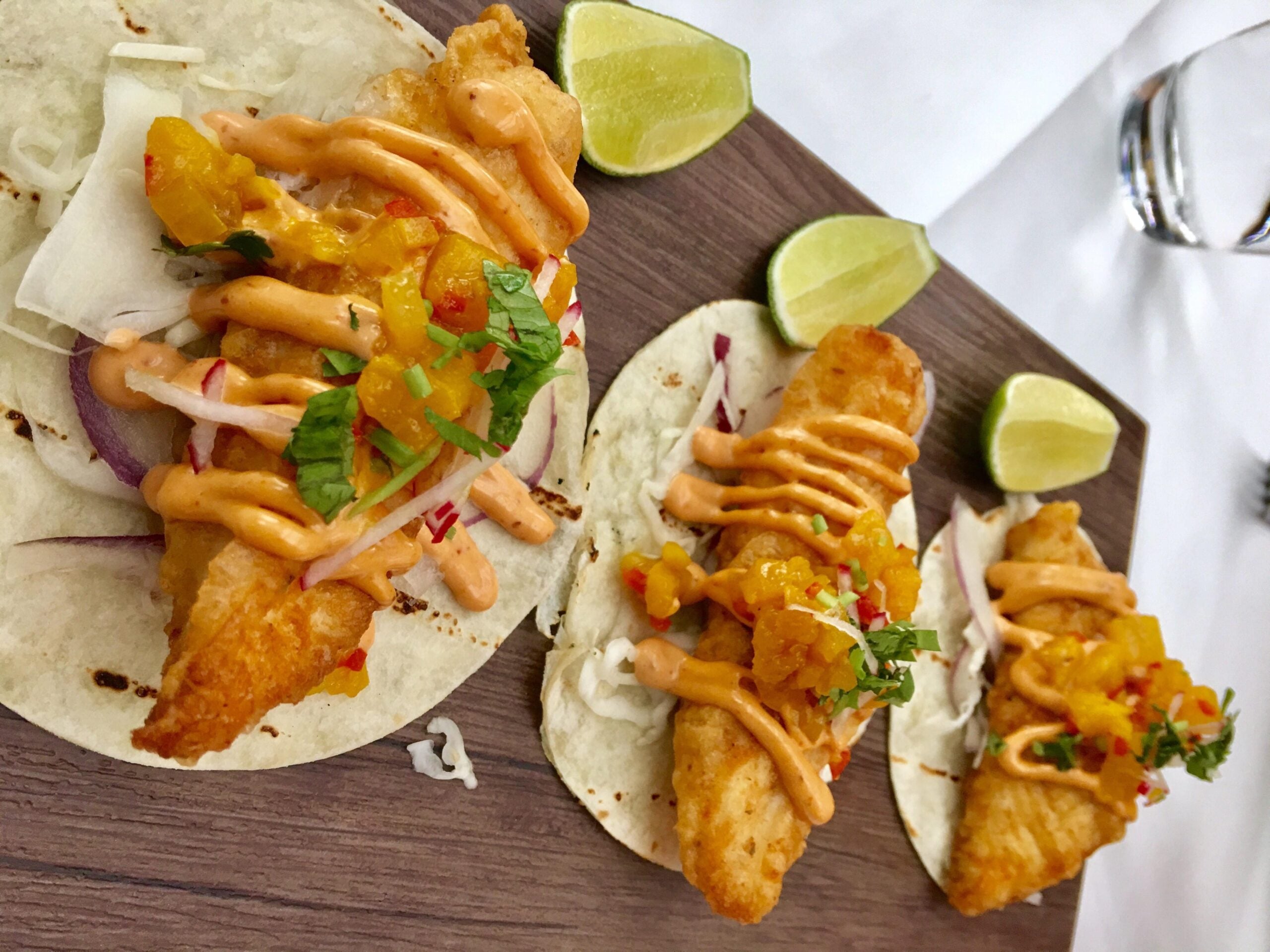 Three fish tacos on flour tortillas, topped with mango salsa, cilantro, and chipotle sauce, served with lime wedges on a wooden board.