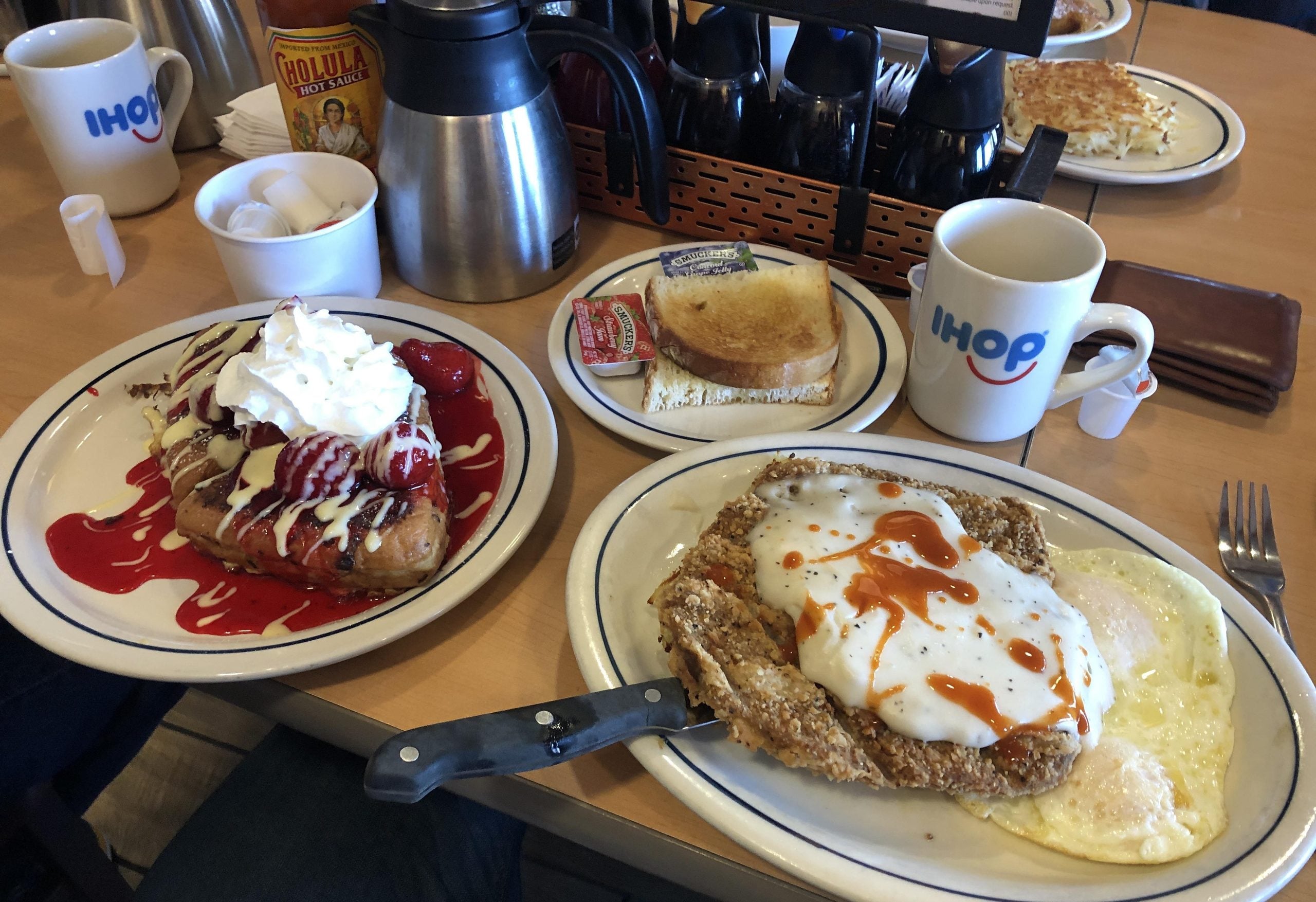 A breakfast spread at IHOP featuring strawberry topped French toast, country fried steak with eggs, toast, and two mugs of coffee.