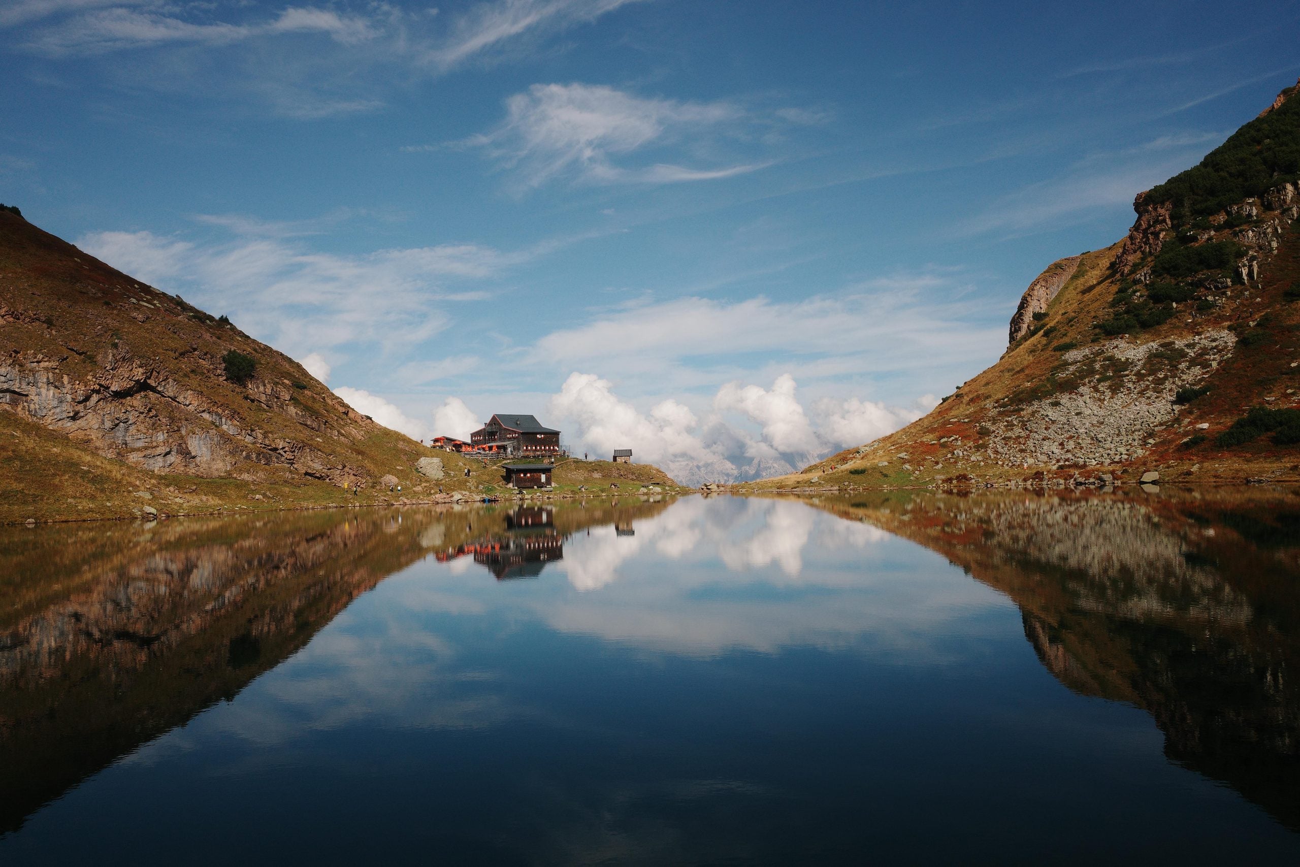 A mountain lake reflects the surrounding grassy slopes and a small cluster of buildings sitting on the distant shore.