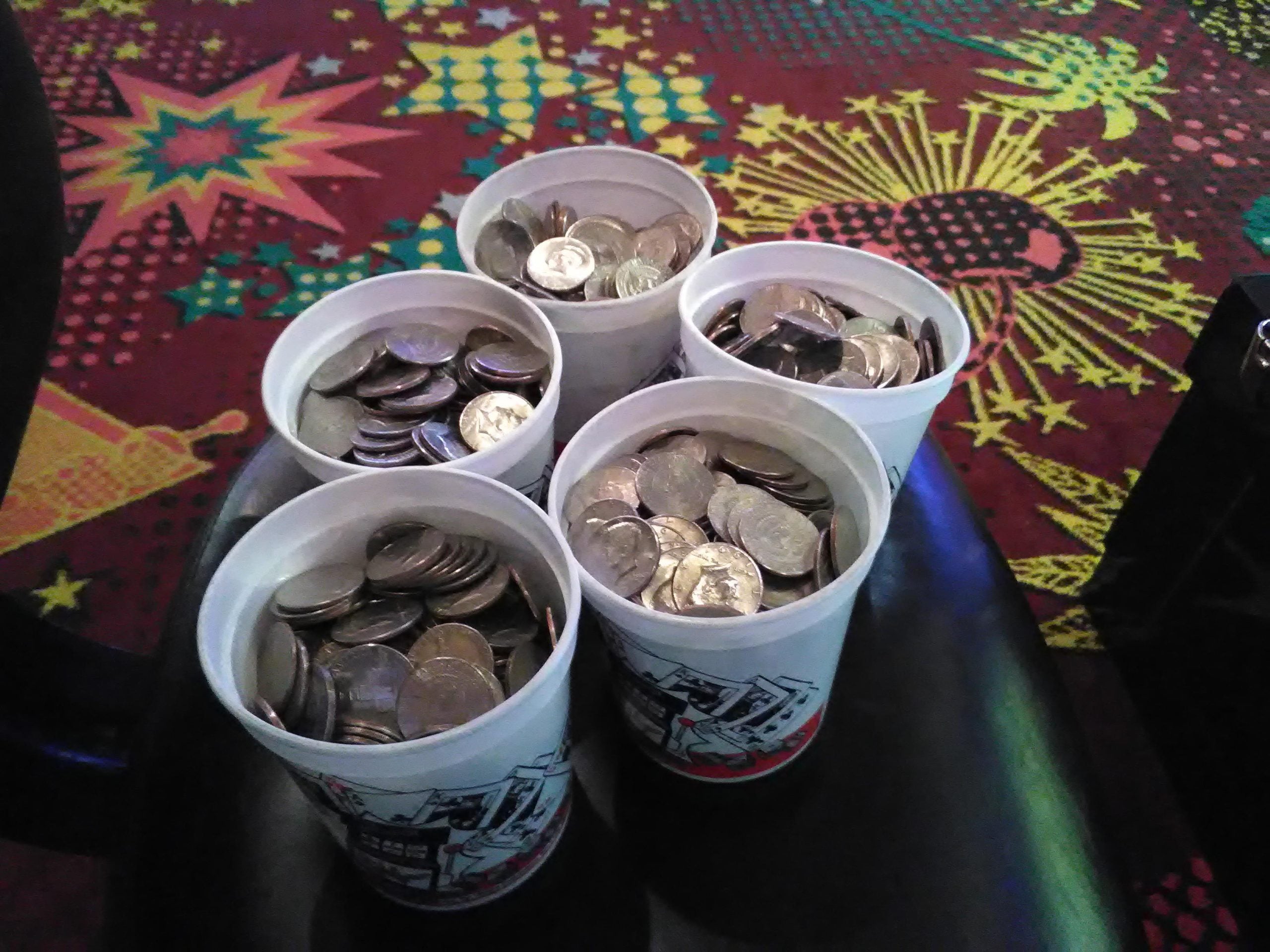 Four white cups filled with quarters and other coins sit on a black chair, evoking Old Las Vegas Traditions, with a colorful, patterned carpet in the background.