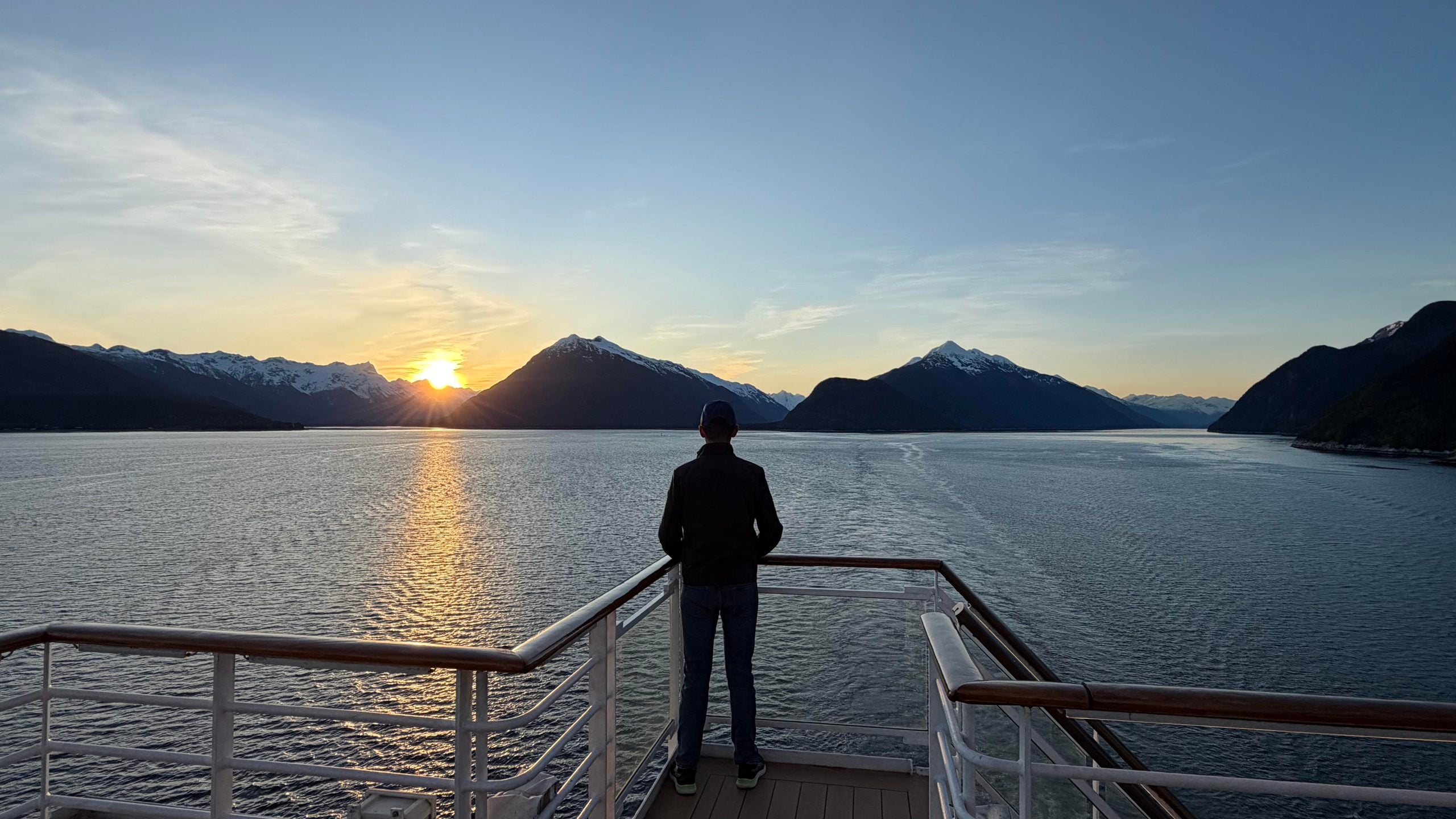 A person stands on the deck of a cruise ship, looking out at a sunset over snow-capped mountains and water.