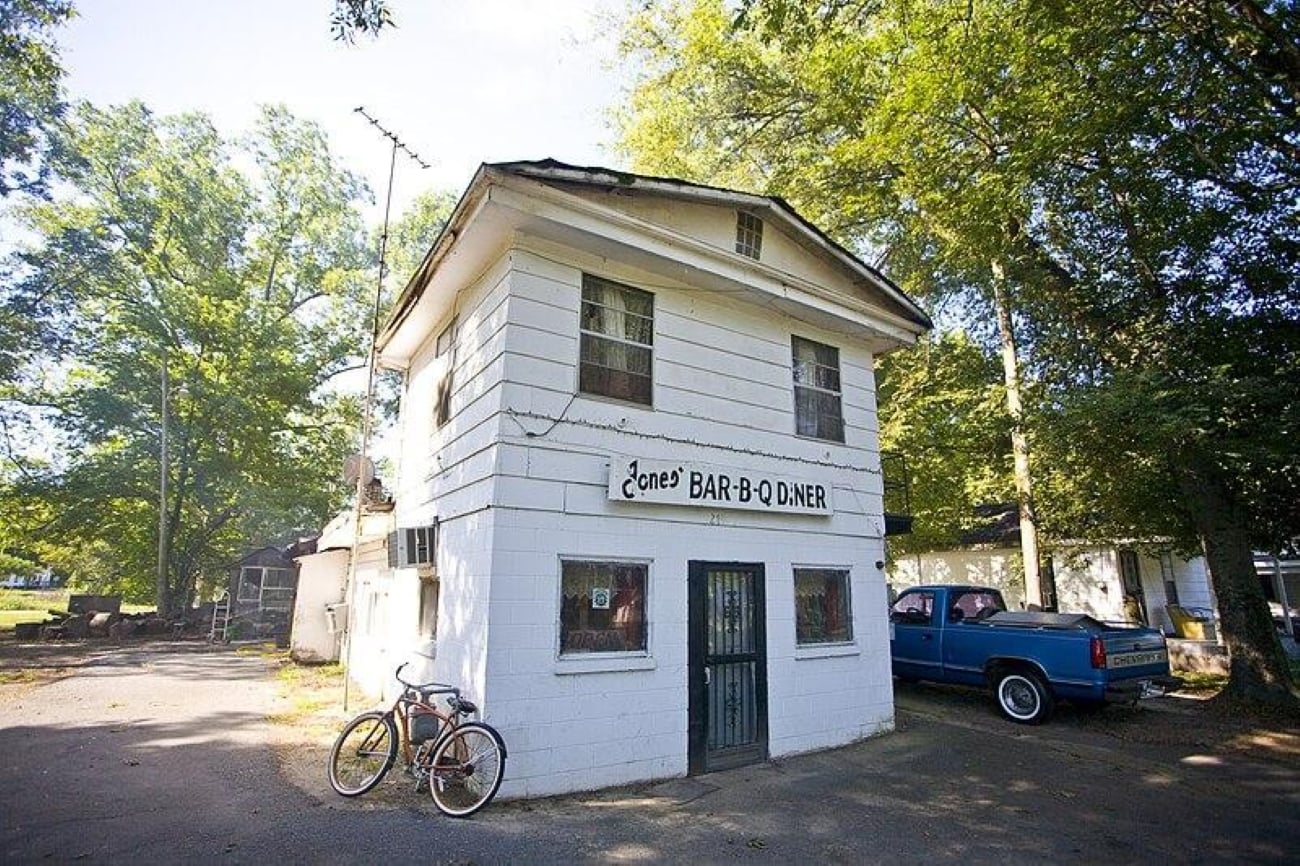 A two-story white building labeled “Jones Bar-B-Q Diner” with a bicycle parked out front and a blue pickup truck parked to the side.