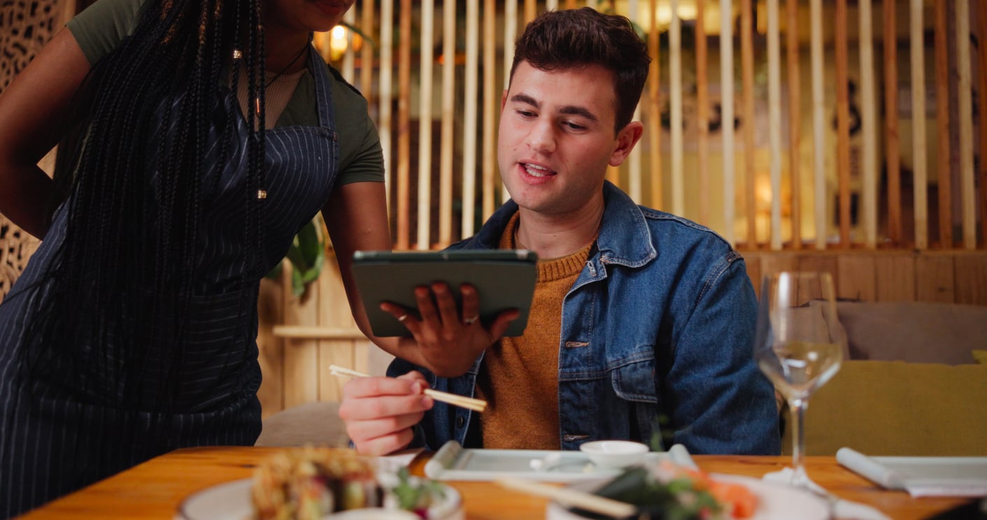 A server in an apron shows a menu on a digital tablet to a young man sitting at a restaurant table with sushi.