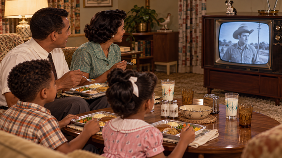 A family eats dinner from TV trays in a living room while watching a cowboy movie on a vintage console television.
