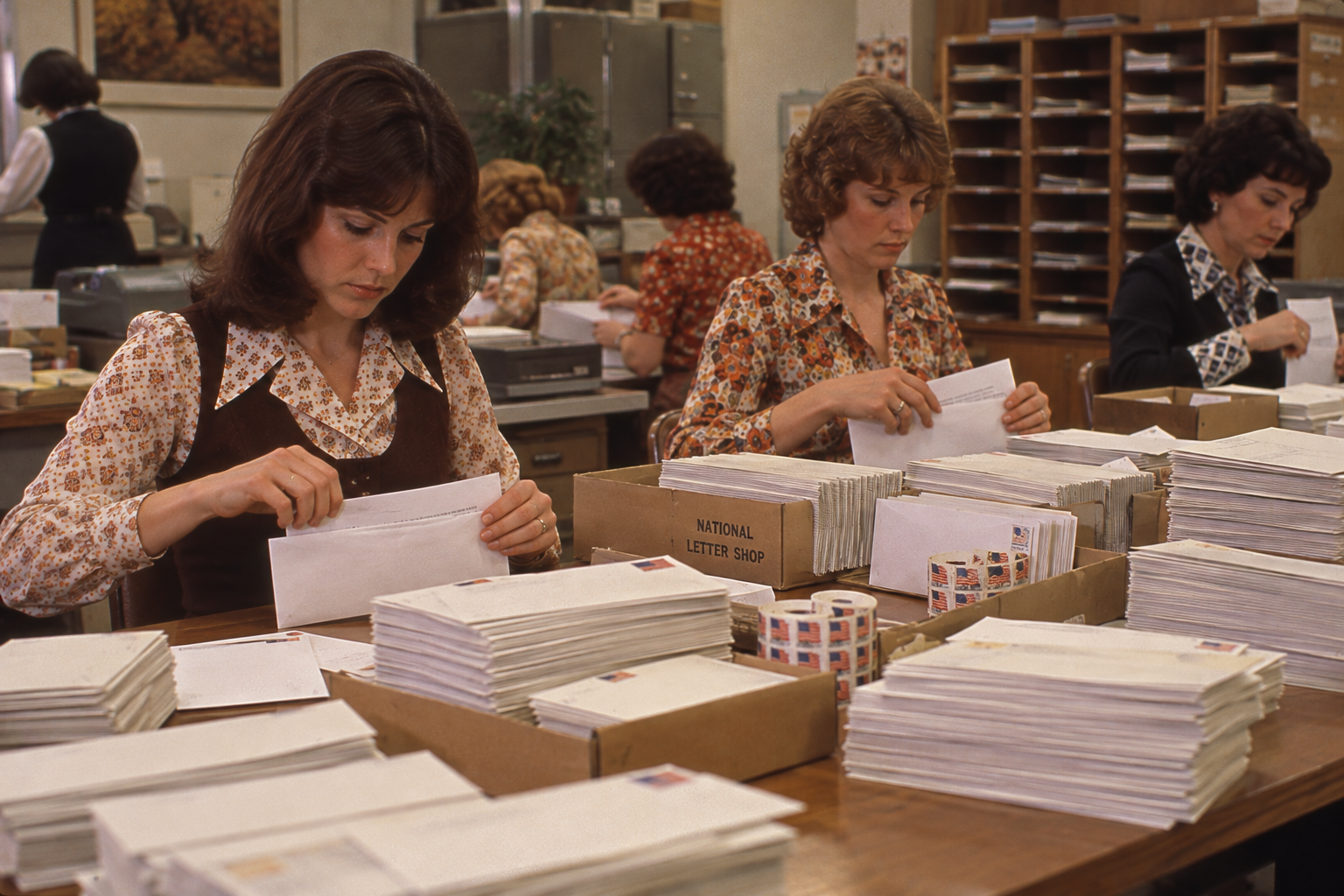 Four women work at desks in an office, processing and sorting large stacks of envelopes.