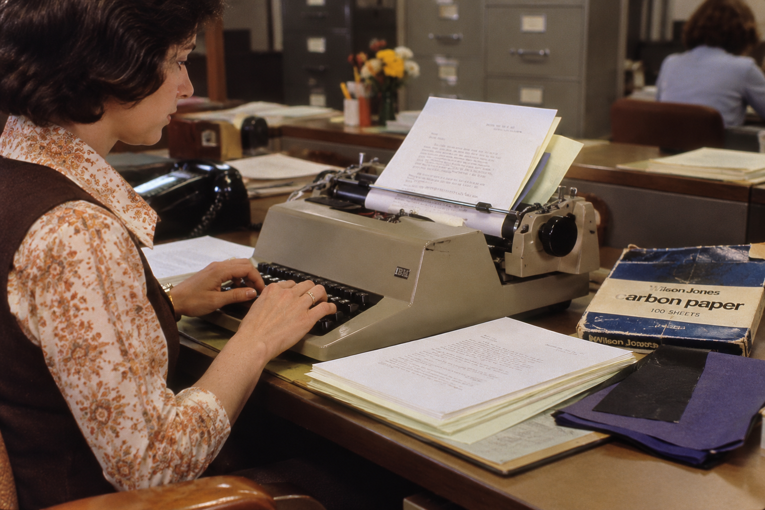 A woman types on an IBM Selectric typewriter at a desk next to a box of Wilson Jones carbon paper.