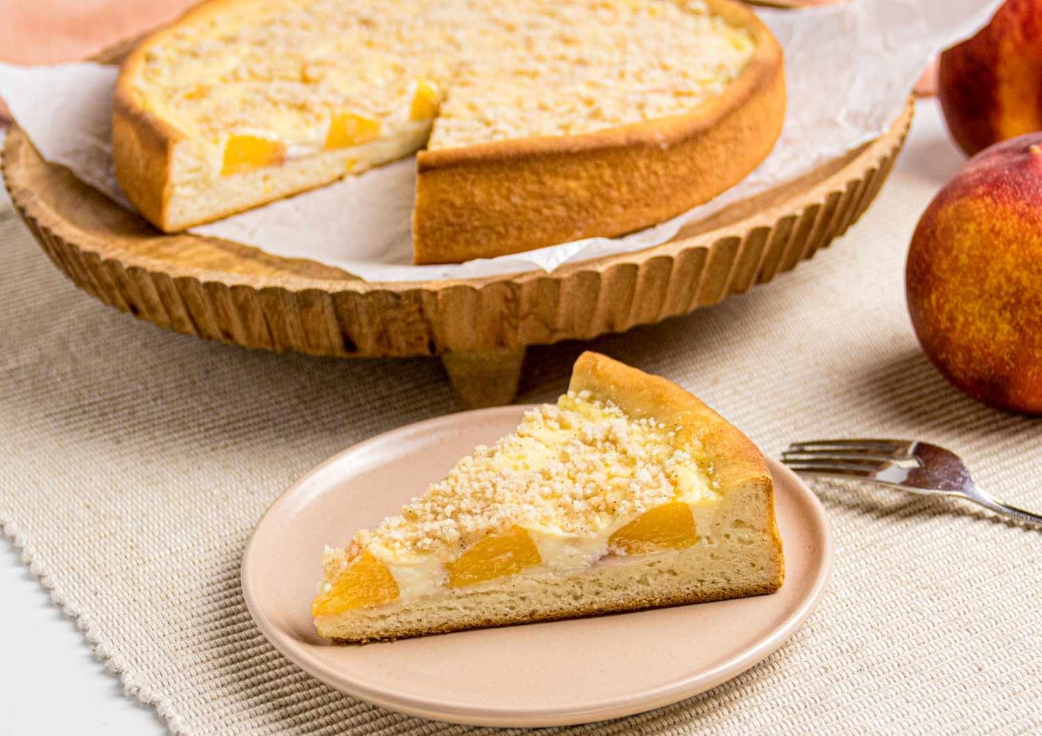A slice of regional dessert peach crumble tart on a small plate, with the rest of the tart on a wooden pedestal and two peaches nearby.
