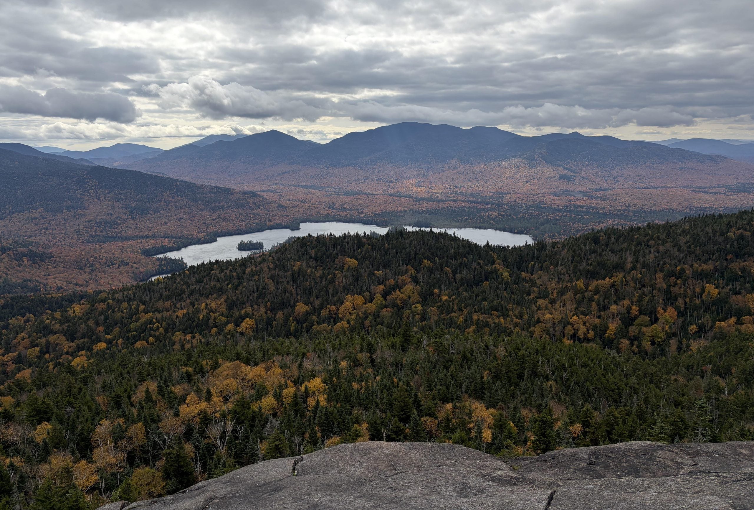 High-angle view of a lake nestled among forested mountains during autumn, seen from a rocky peak under a cloudy sky.