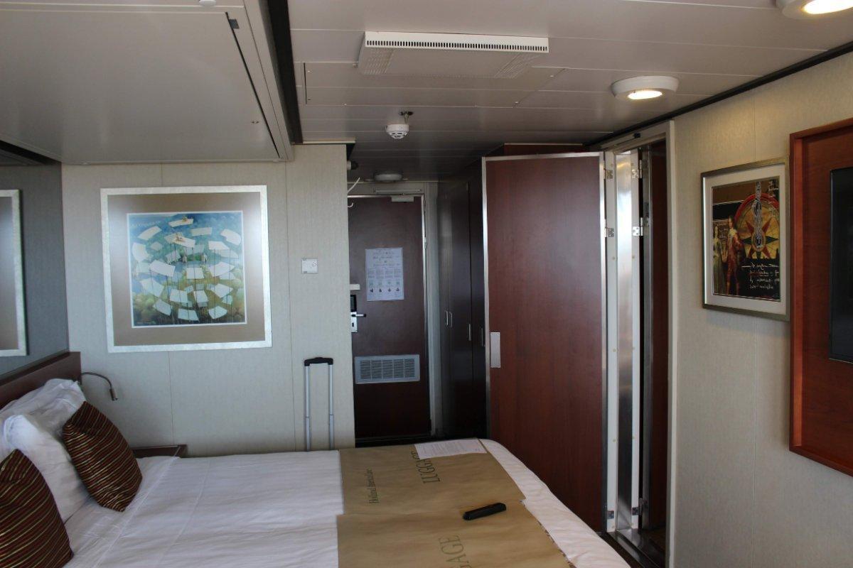 Interior of a cruise ship cabin with a bed, luggage tags on the bedspread, and a doorway leading to a small hallway.