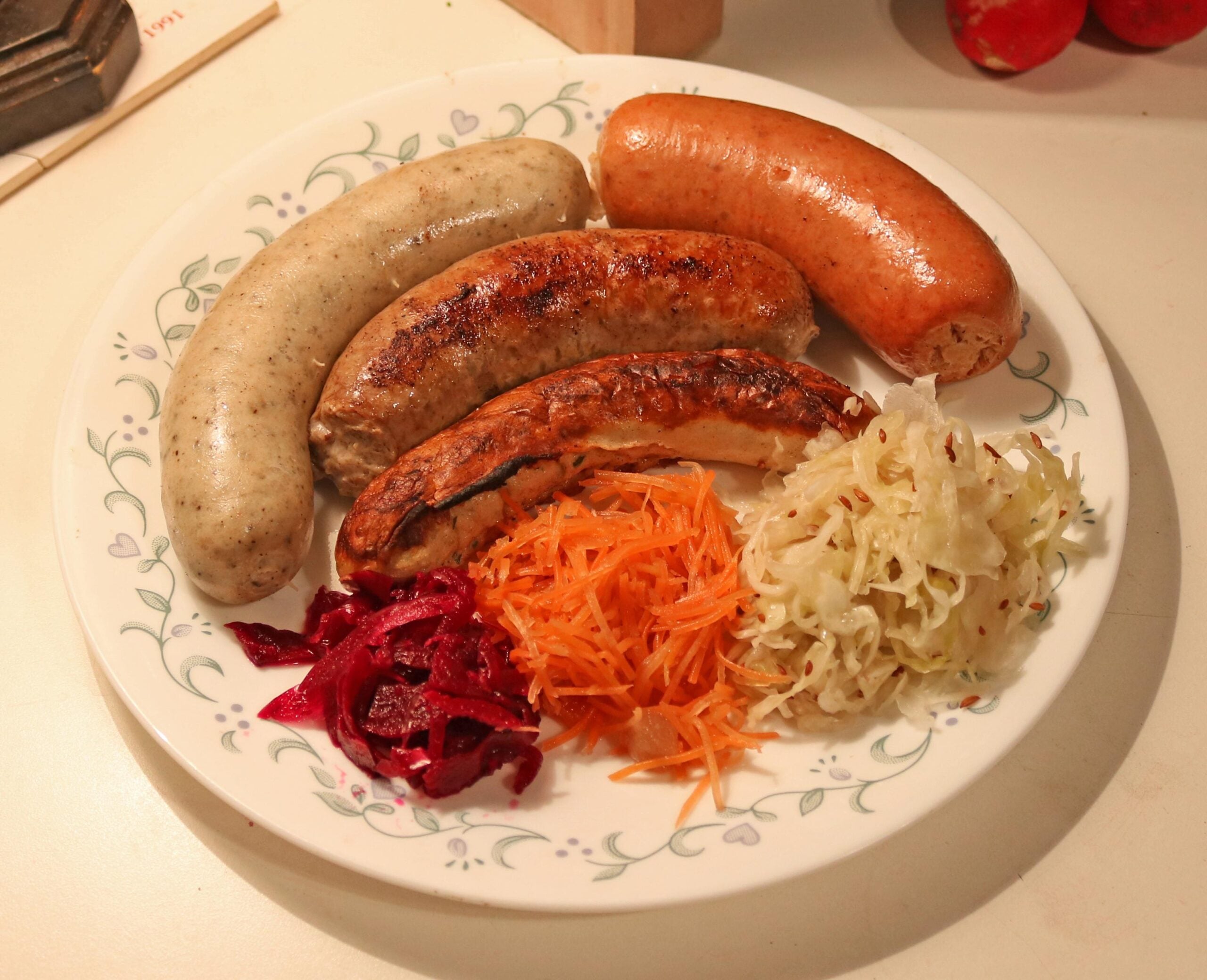 A plate with three sausages alongside small piles of shredded beets, carrots, and sauerkraut.
