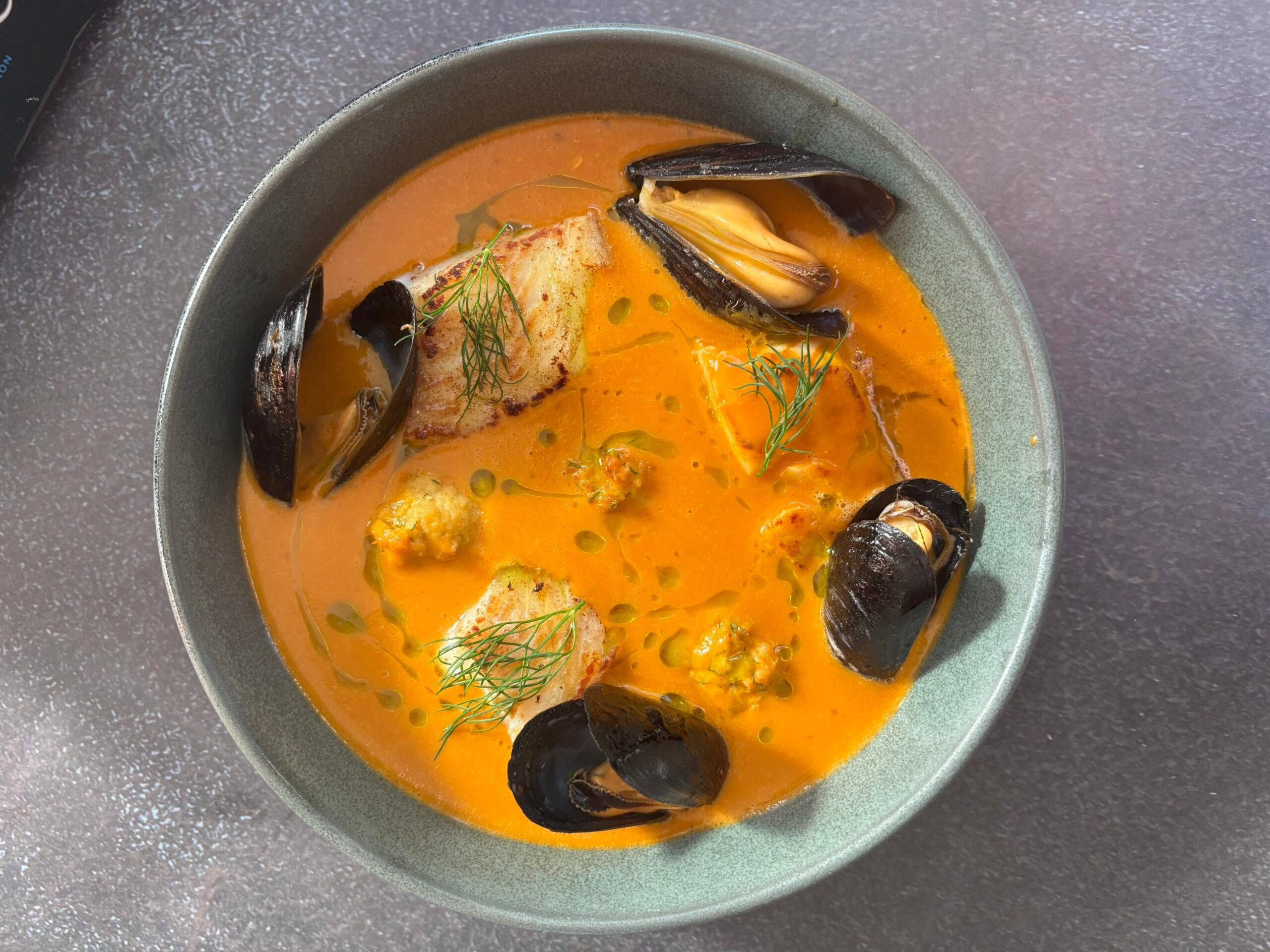 A bowl of orange seafood stew garnished with mussels, pieces of white fish, and sprigs of dill.