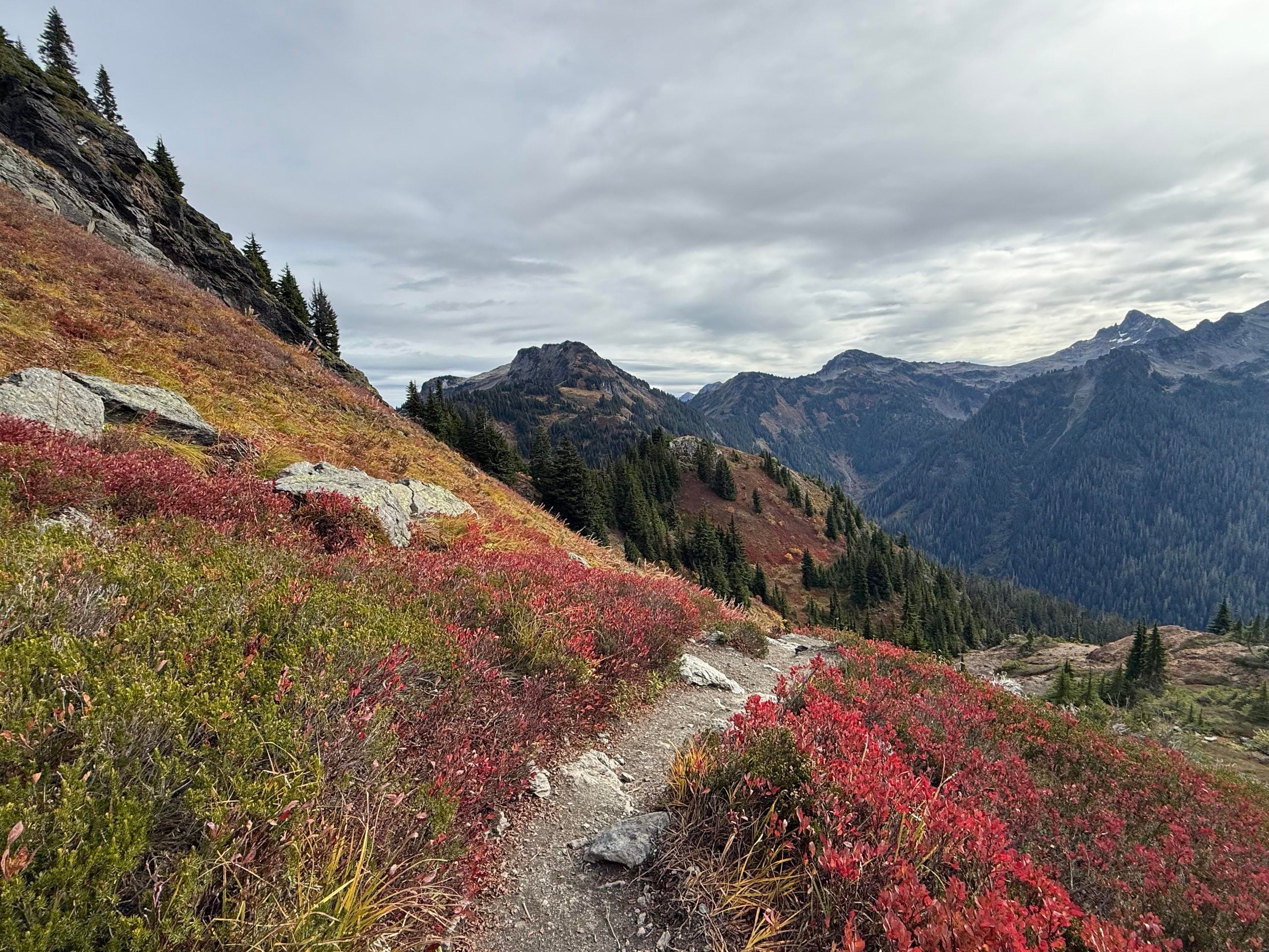 A winding dirt hiking trail through a mountain landscape covered in red and orange autumn foliage under a cloudy sky.