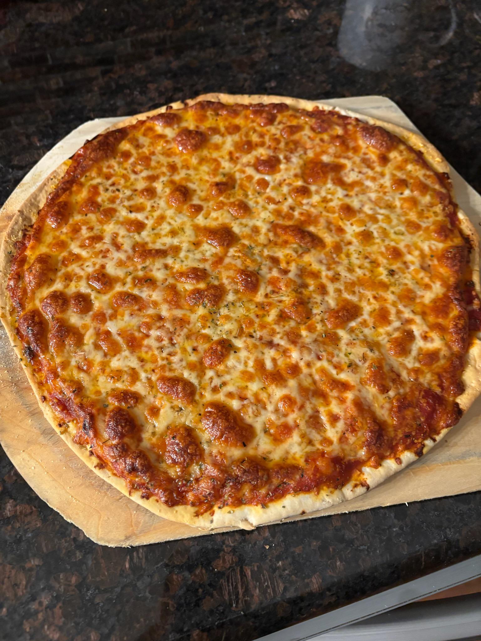 A whole cheese pizza with a golden, bubbly crust and melted cheese, sitting on a wooden pizza peel atop a dark kitchen countertop.