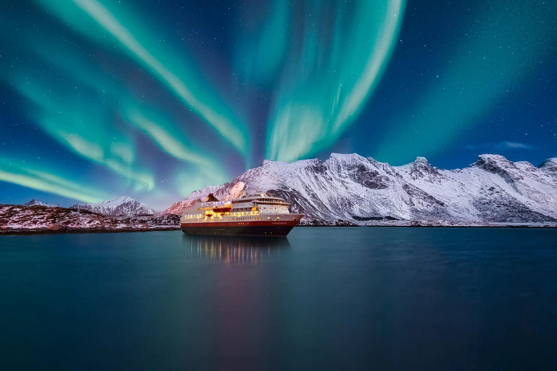 A cruise ship anchored in a calm bay at night under vibrant green northern lights and snow-covered mountains.