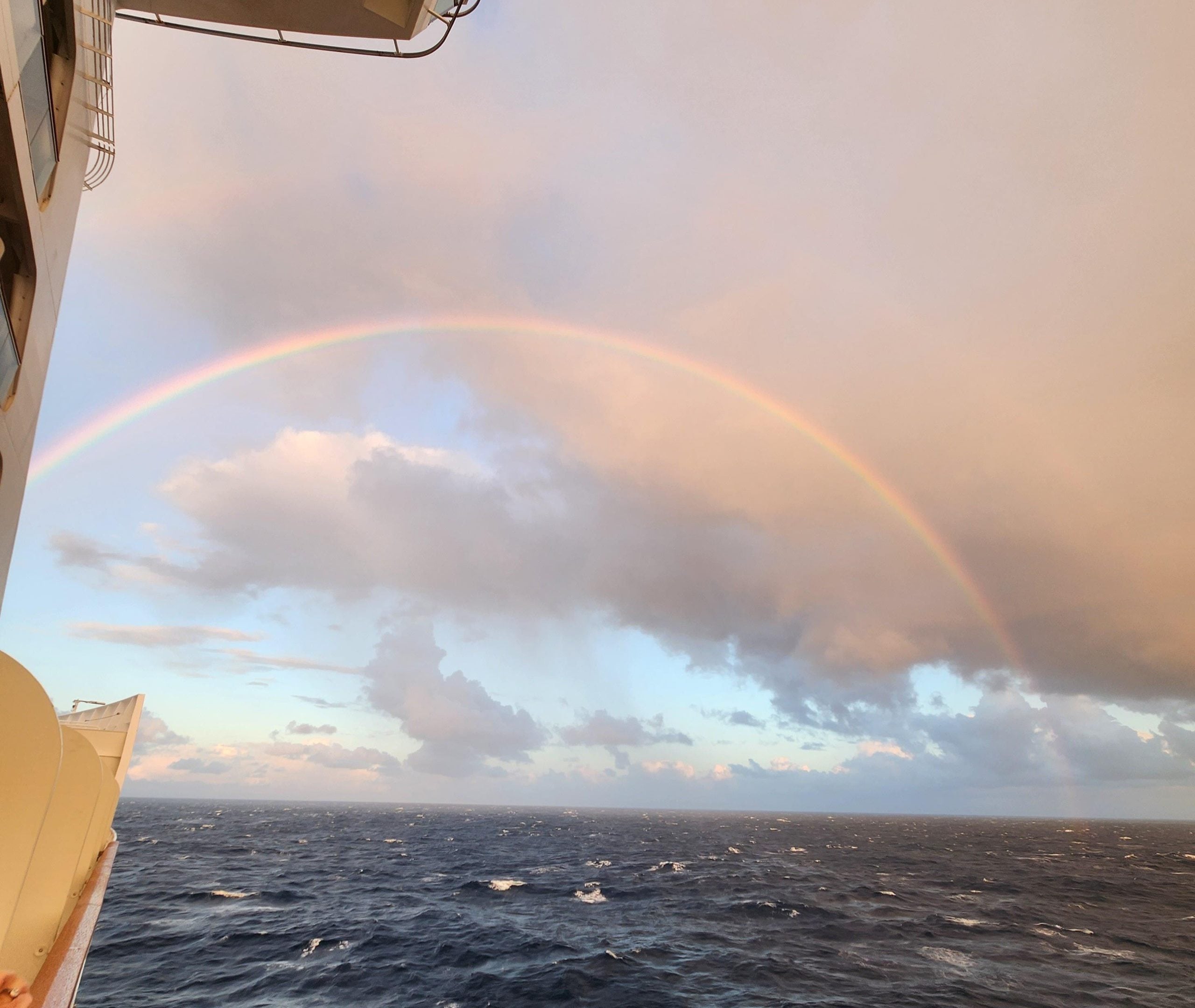 A rainbow arches over the open ocean, viewed from the side of a cruise ship.