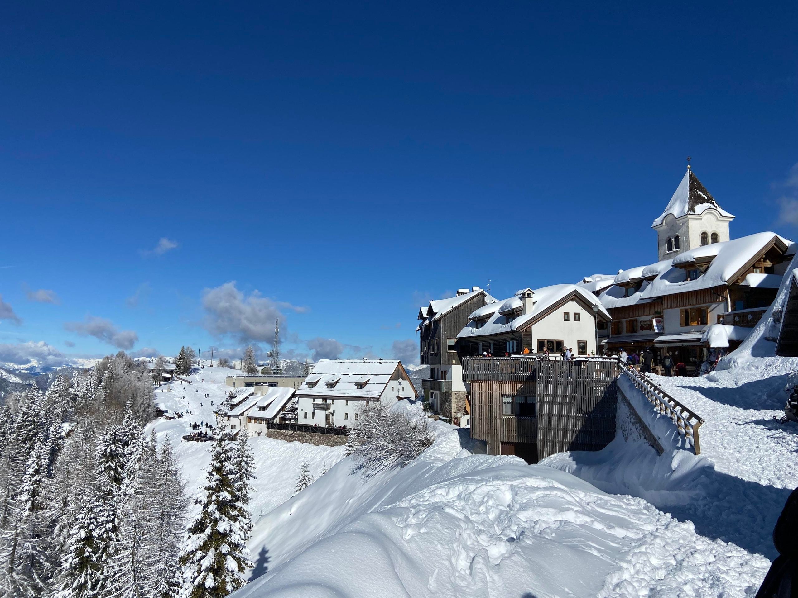 Snow-covered mountain buildings and a small church nestled on a hillside against a clear blue sky.