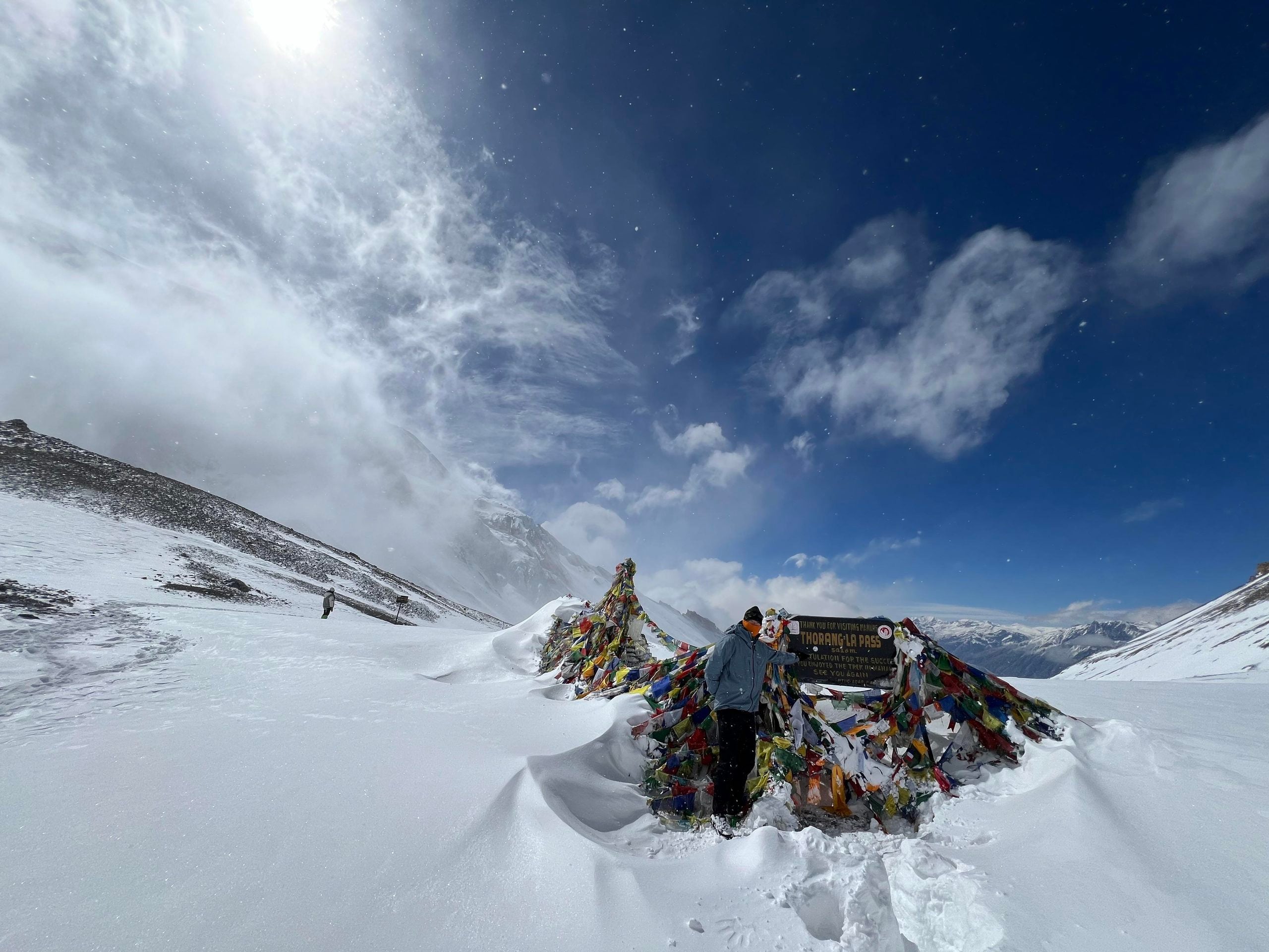 A hiker stands by a sign marking the Thorong La Pass, surrounded by snow and colorful prayer flags under a bright sky.