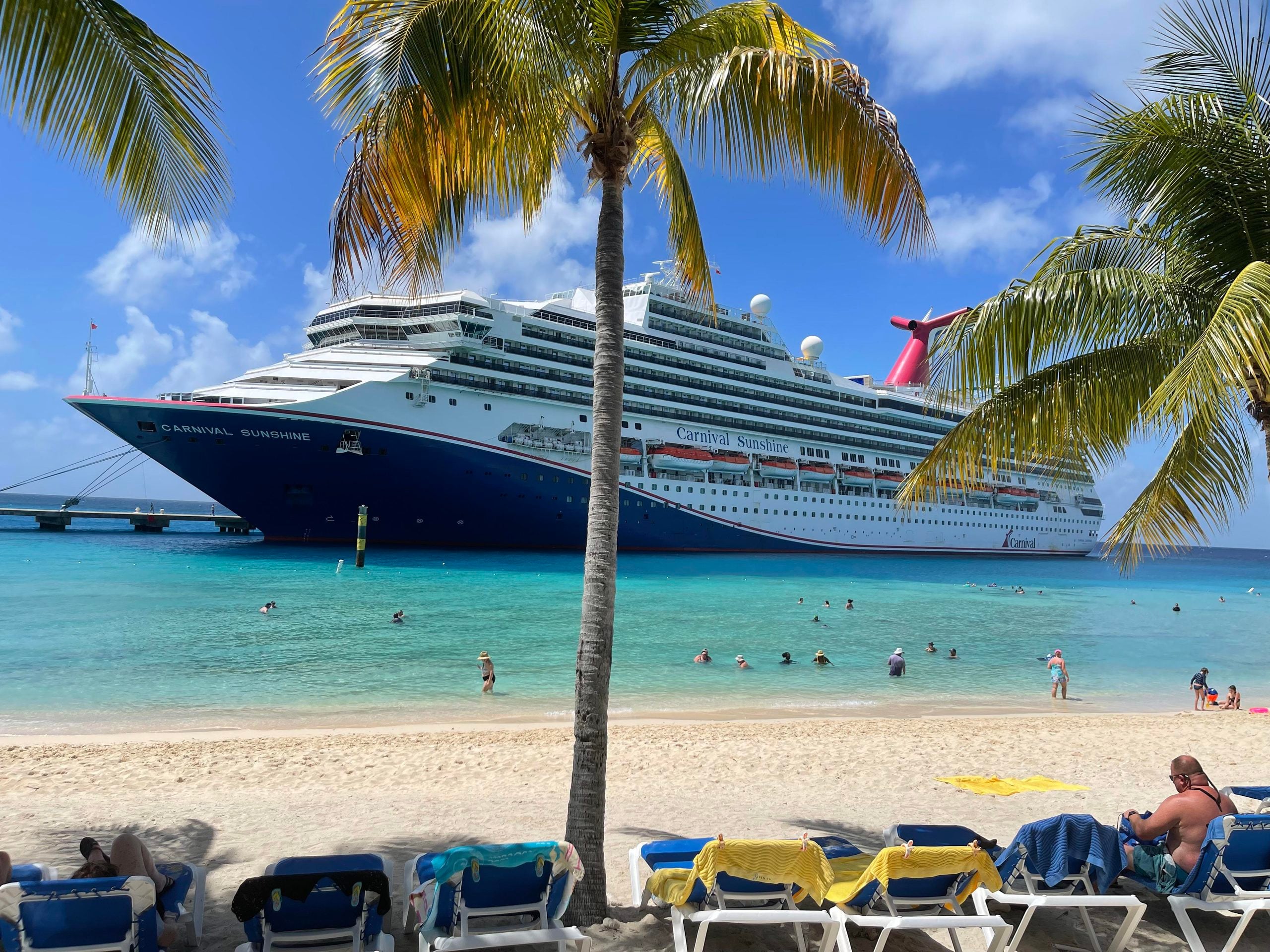 A Carnival Sunshine cruise ship docked near a tropical beach, where people swim in turquoise water alongside rows of lounge chairs on the sand, captured in these cruise ship photos.