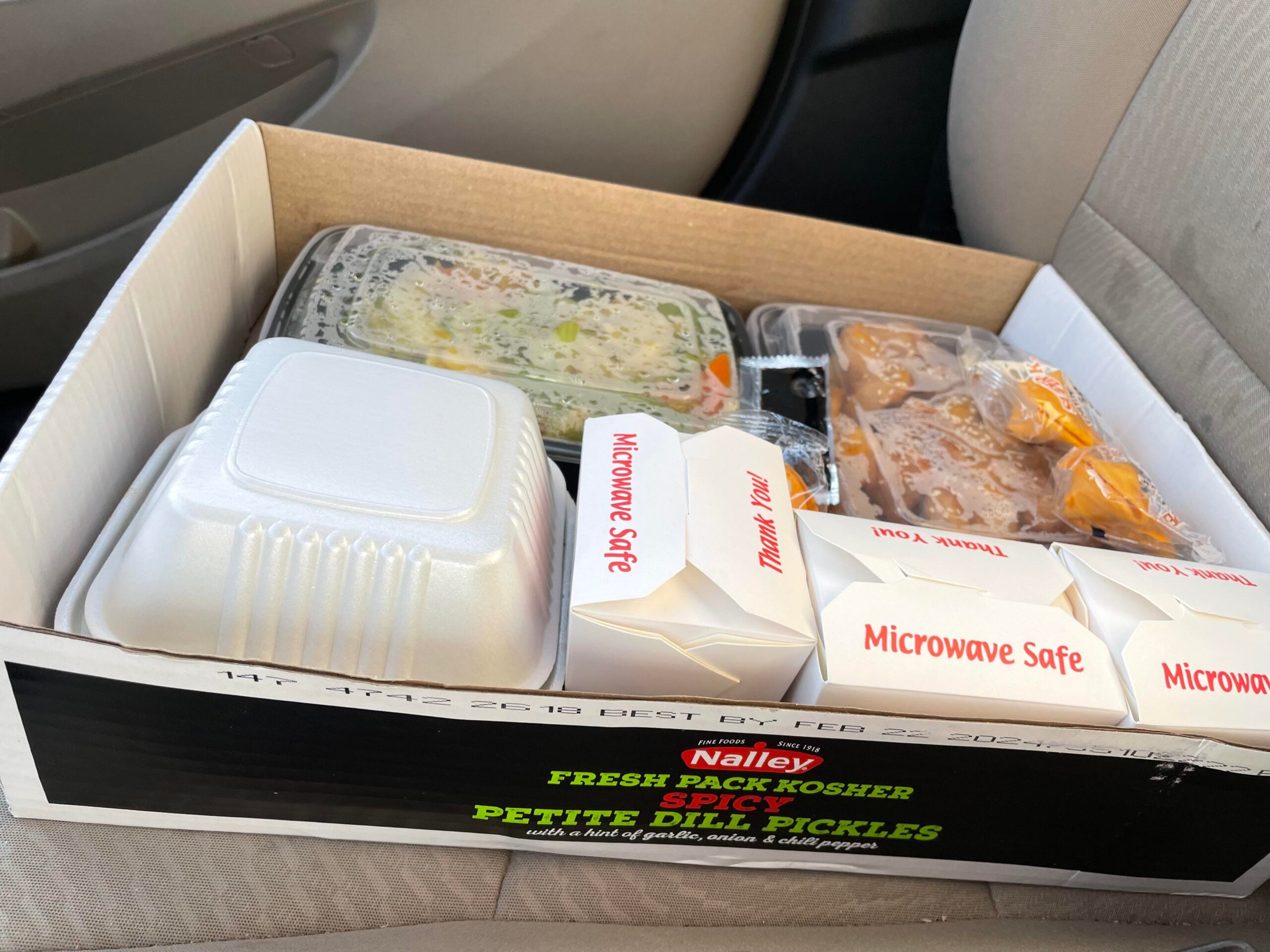 A cardboard box containing several food containers, including two small white boxes labeled “Microwave Safe,” sitting on a car seat.