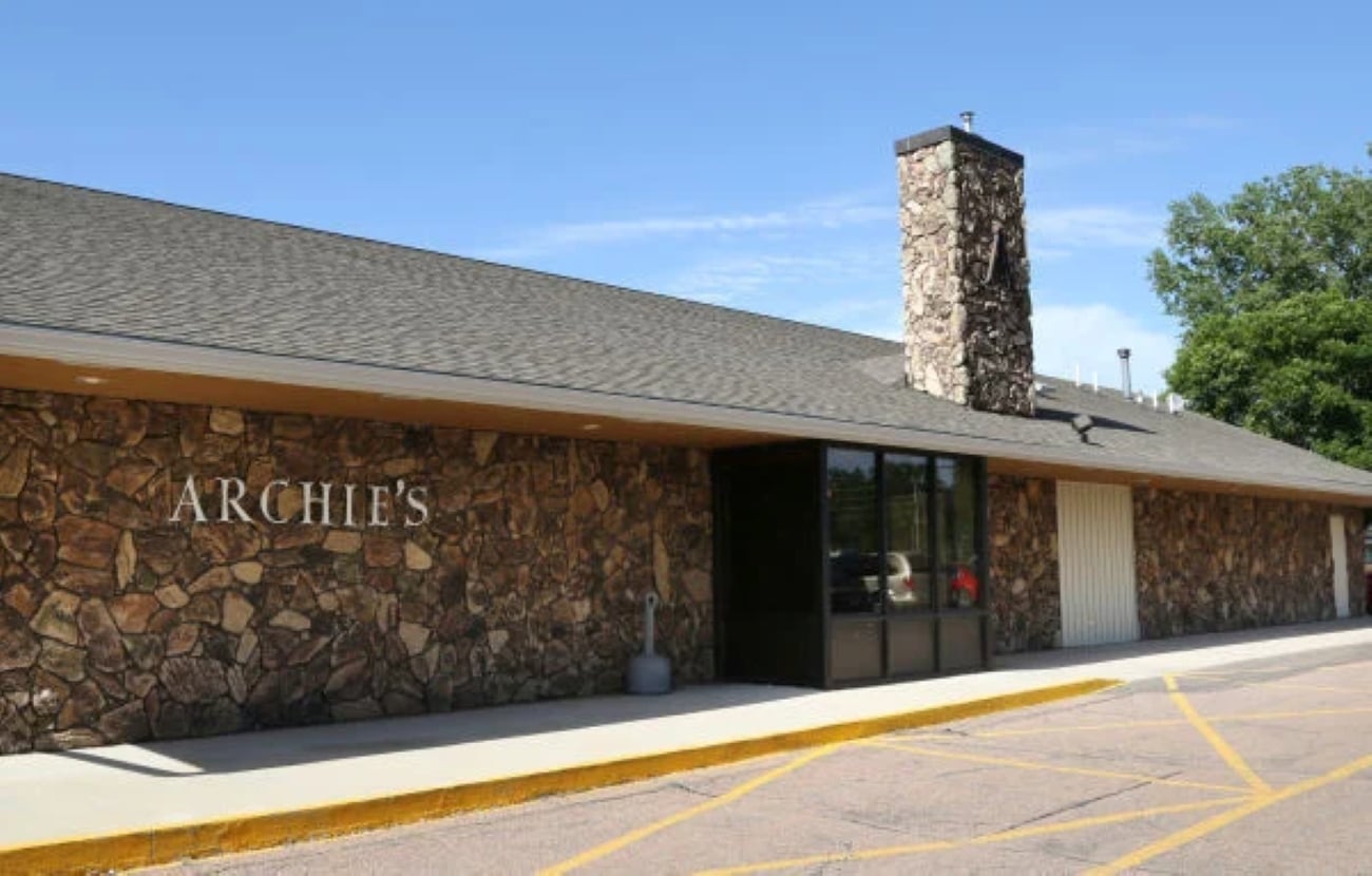 The exterior of Archie’s restaurant, featuring stone walls, a large stone chimney, and a paved parking lot.