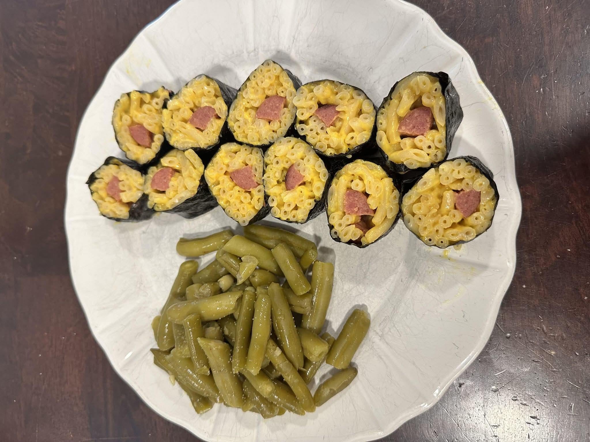Sushi-style rolls made with macaroni and cheese and sliced sausage, served with a side of green beans on a white plate.