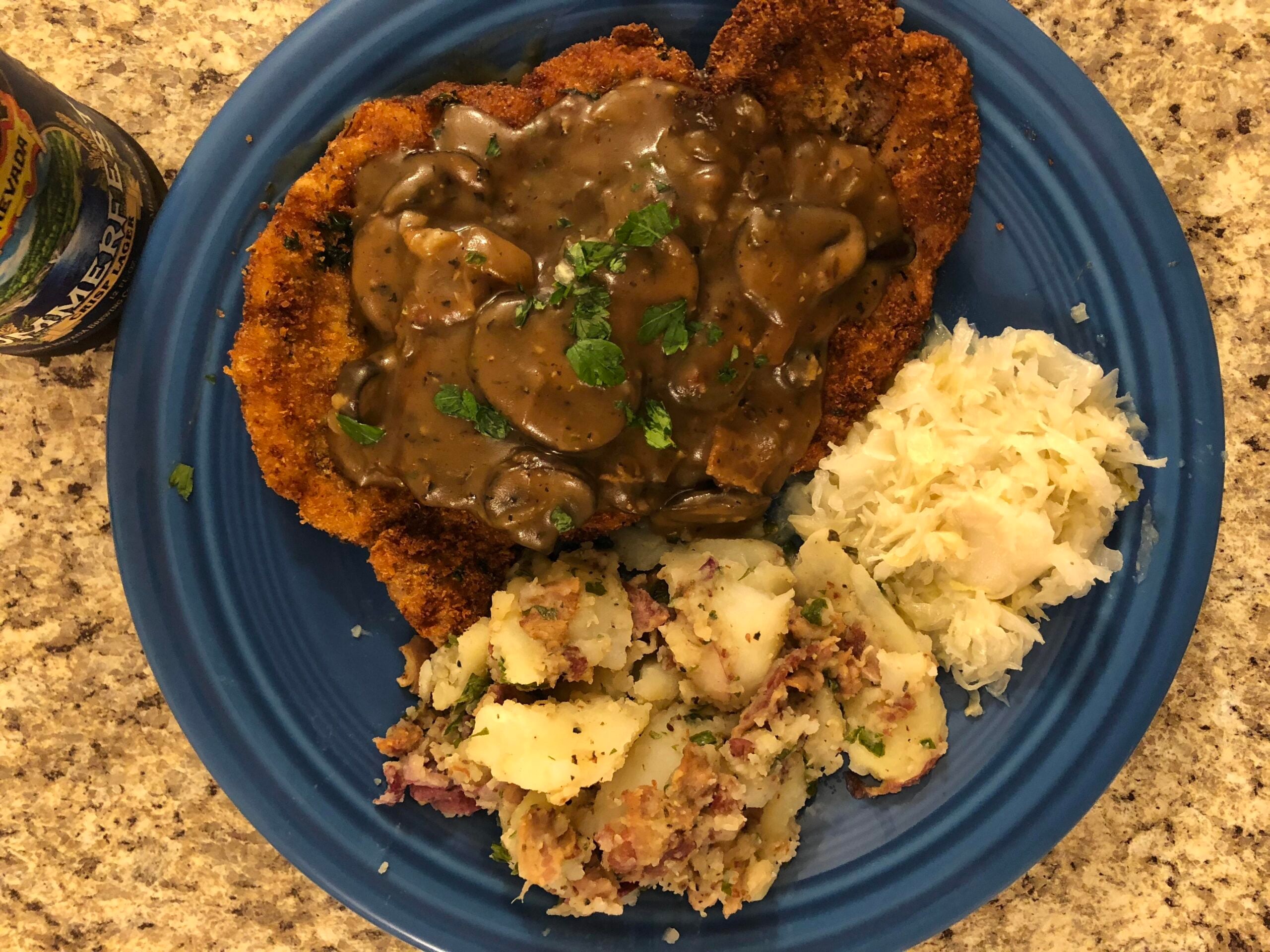 Breaded schnitzel topped with mushroom gravy, served with potato salad and sauerkraut on a blue plate.