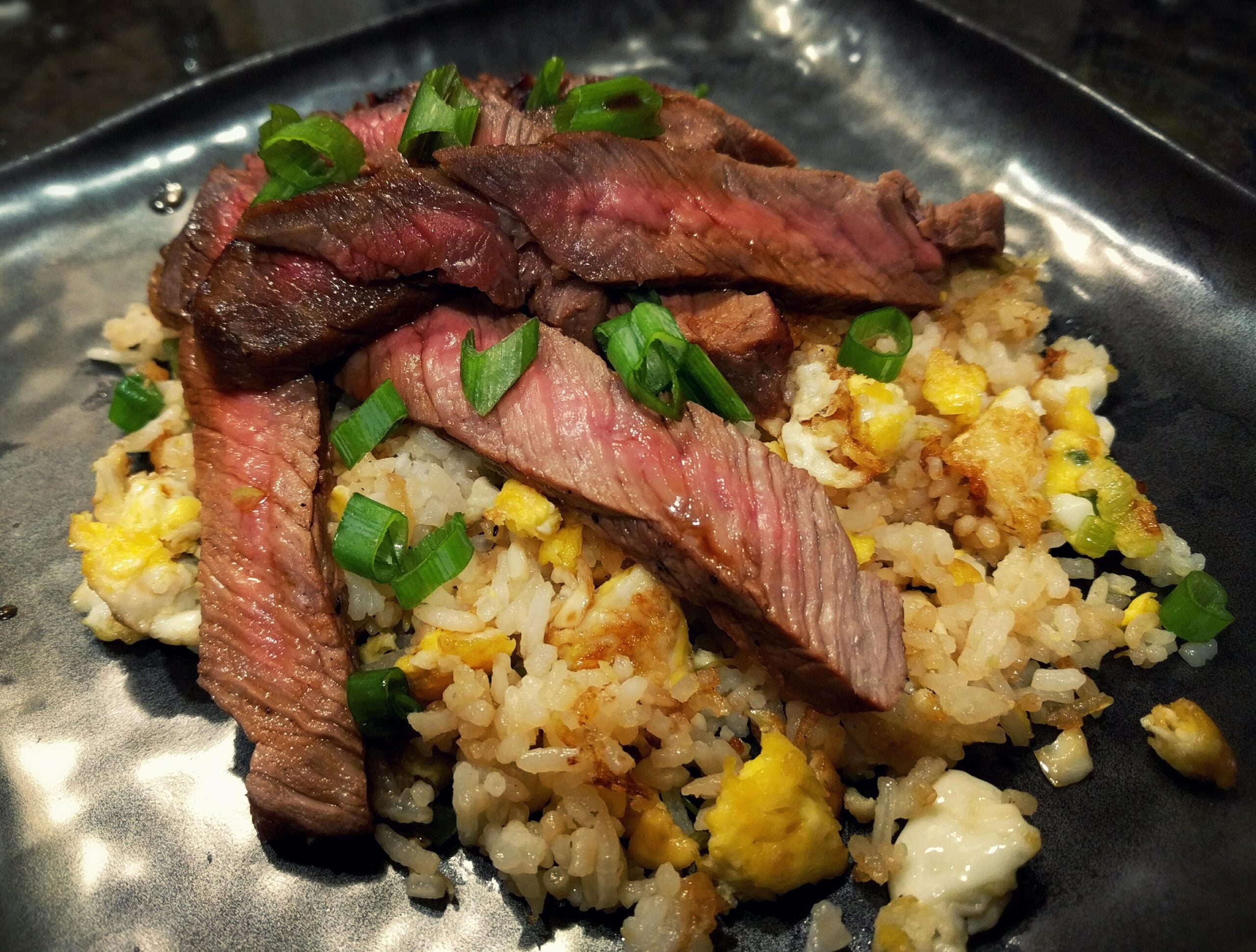 Slices of medium-rare steak over egg fried rice, garnished with green onions.