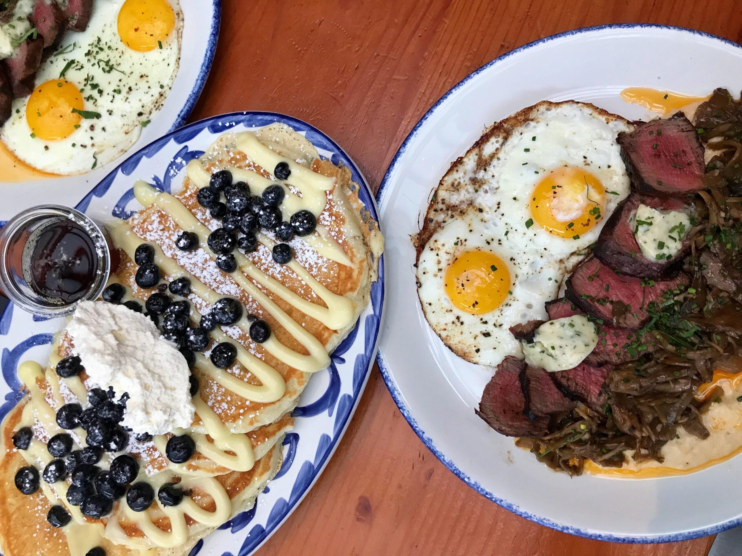A stack of pancakes topped with blueberries and cream, flanked by two plates of steak and sunny-side up eggs on a wooden table.