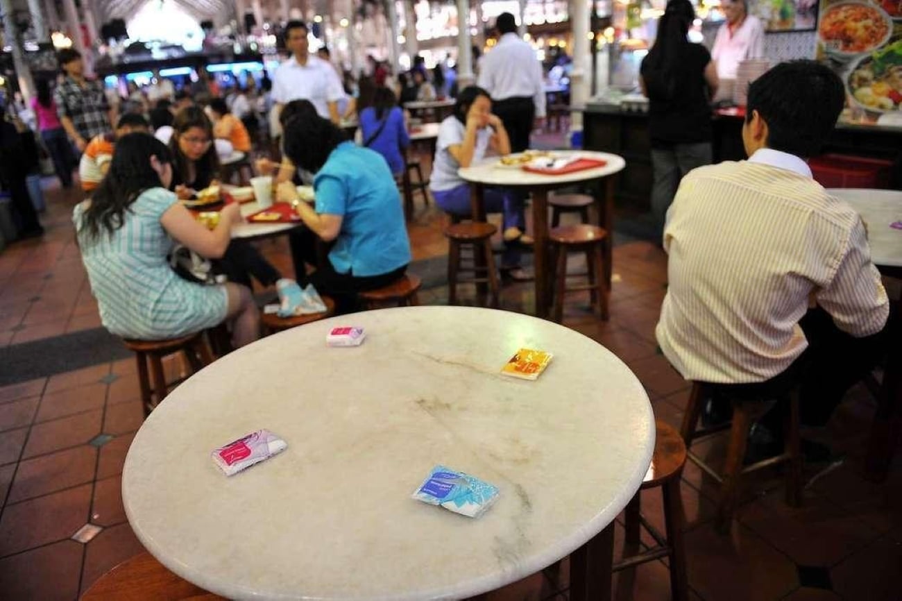 A round marble table in the foreground with four small tissue packets placed on it, with people eating at nearby tables in a bustling hawker center.