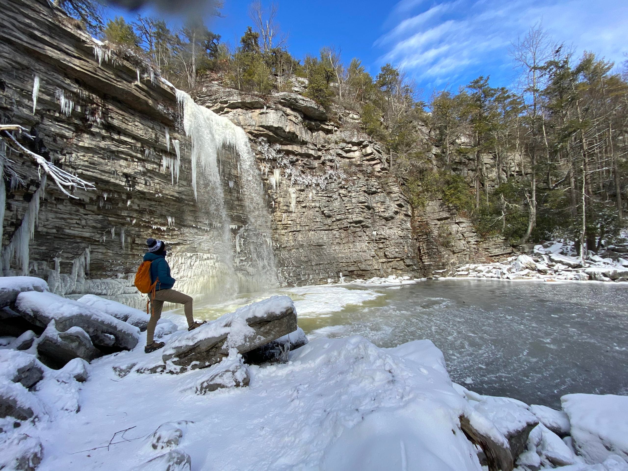 A hiker with an orange backpack stands on a snowy rock looking at a partially frozen waterfall cascading over a cliff.