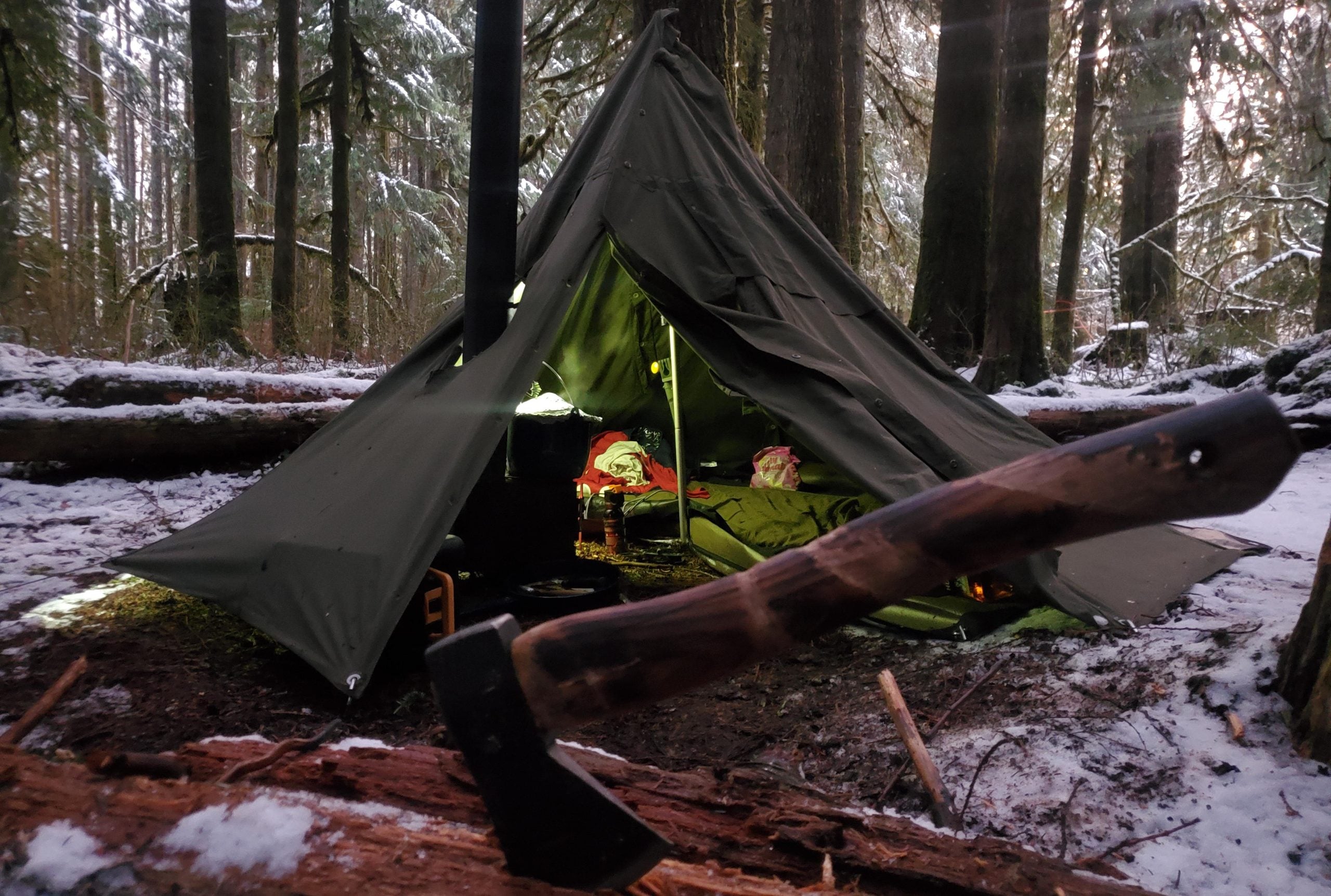 An axe handle rests in the foreground with a green canvas tent pitched in a snowy forest.