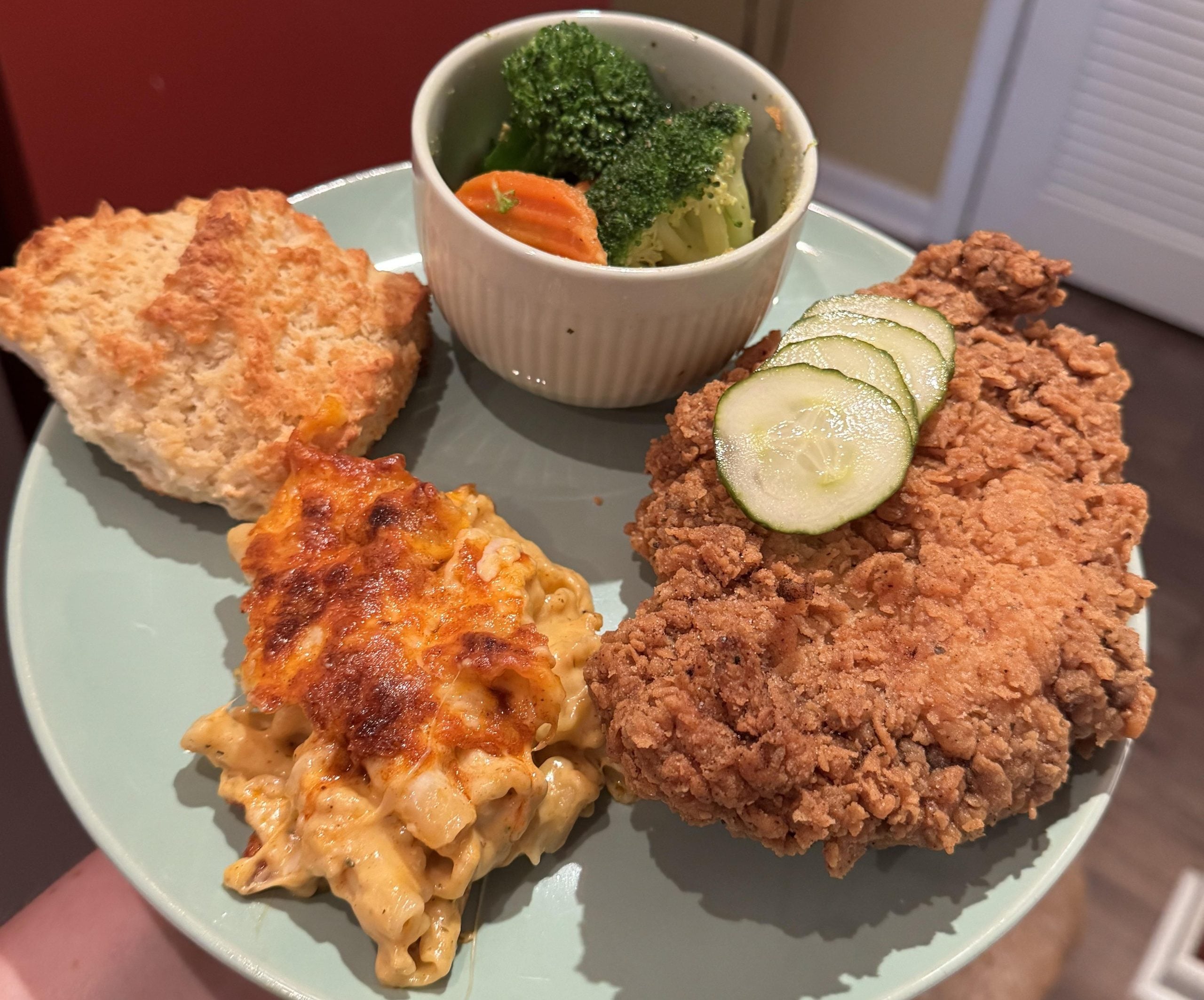 A plate with fried chicken topped with cucumber slices, a serving of baked macaroni and cheese, a biscuit, and a small bowl of broccoli and carrots.