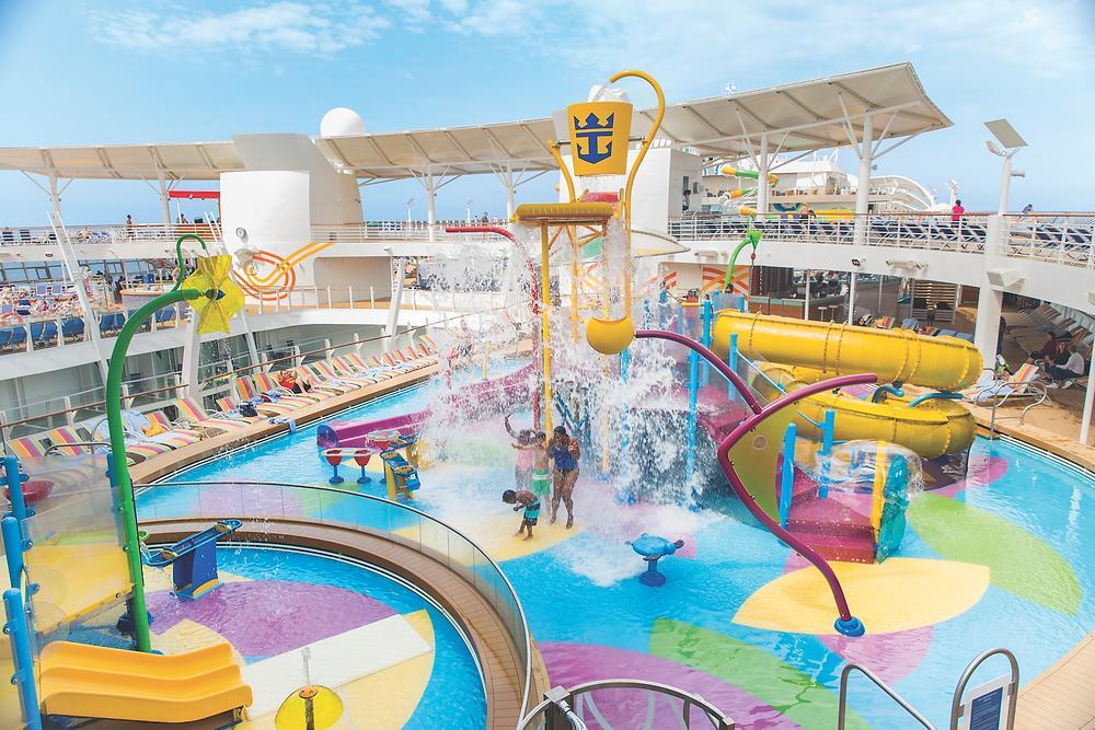 Children play in a colorful cruise ship amenity splash pad area with water slides, spray features, and a large central dumping bucket.