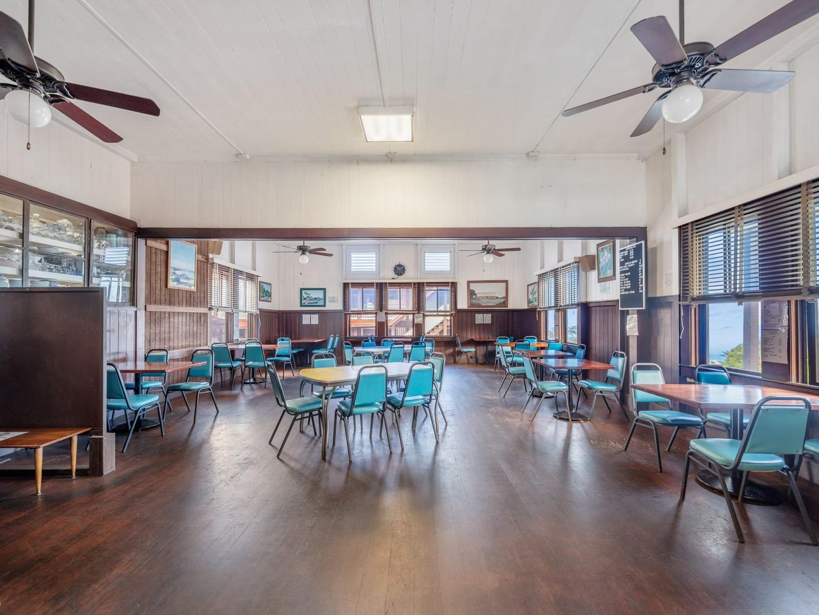 A spacious dining hall with a high ceiling, dark wooden floors, and several tables paired with teal chairs.