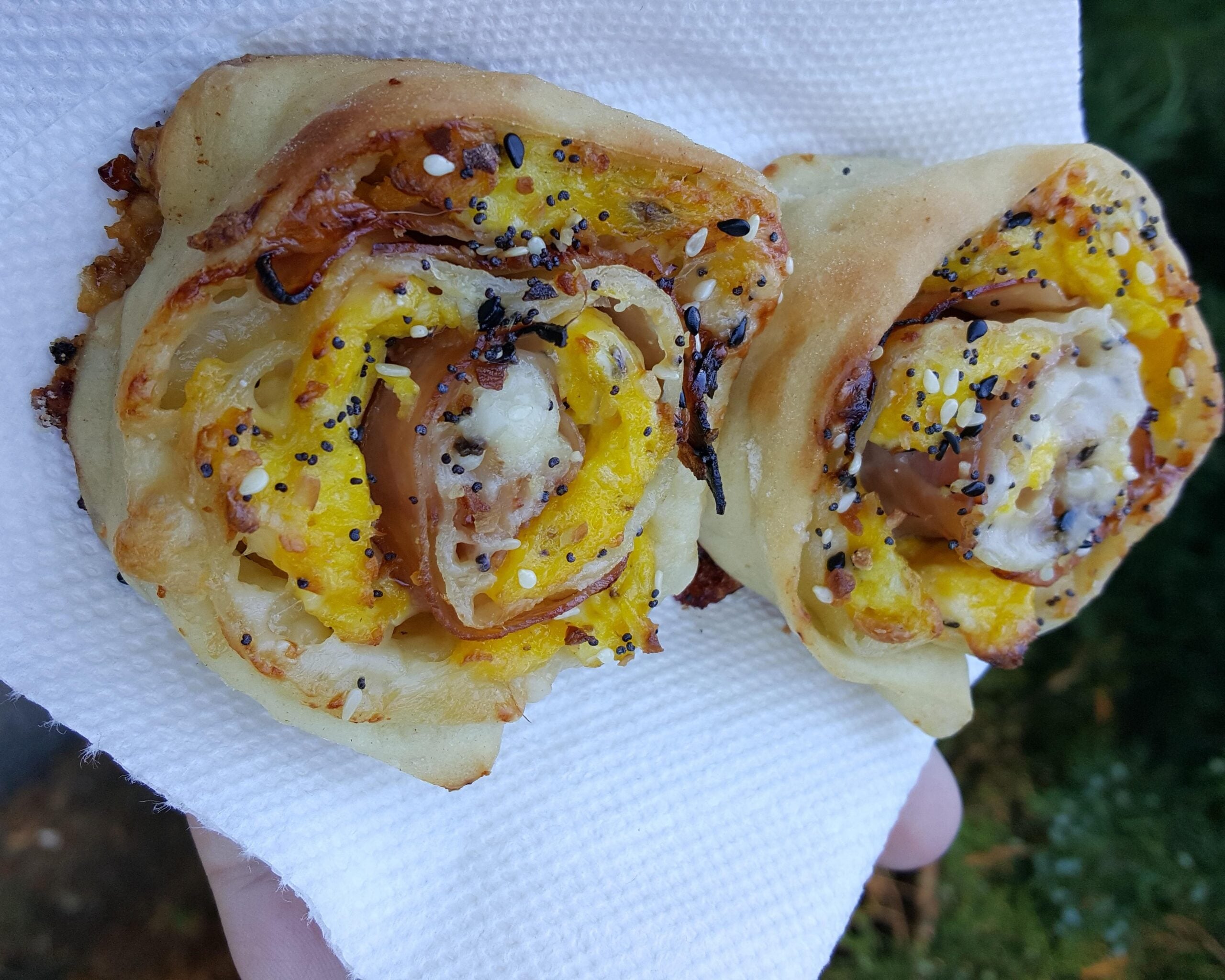 Two rolled breakfast pastries filled with egg, cheese, and ham, topped with everything bagel seasoning on a white napkin.