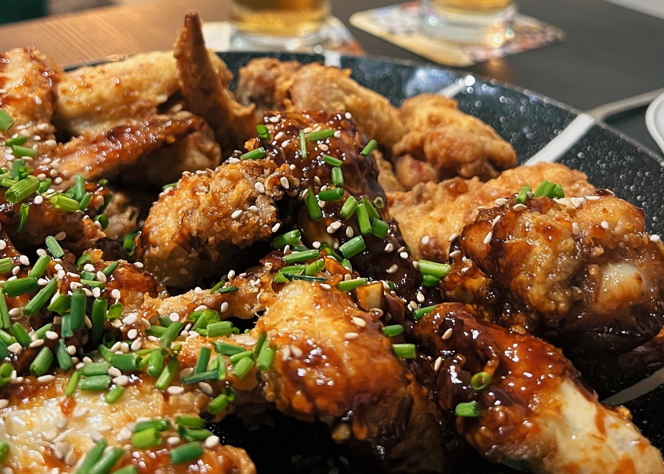 A platter of fried chicken wings topped with a dark glaze, sesame seeds, and chopped chives.