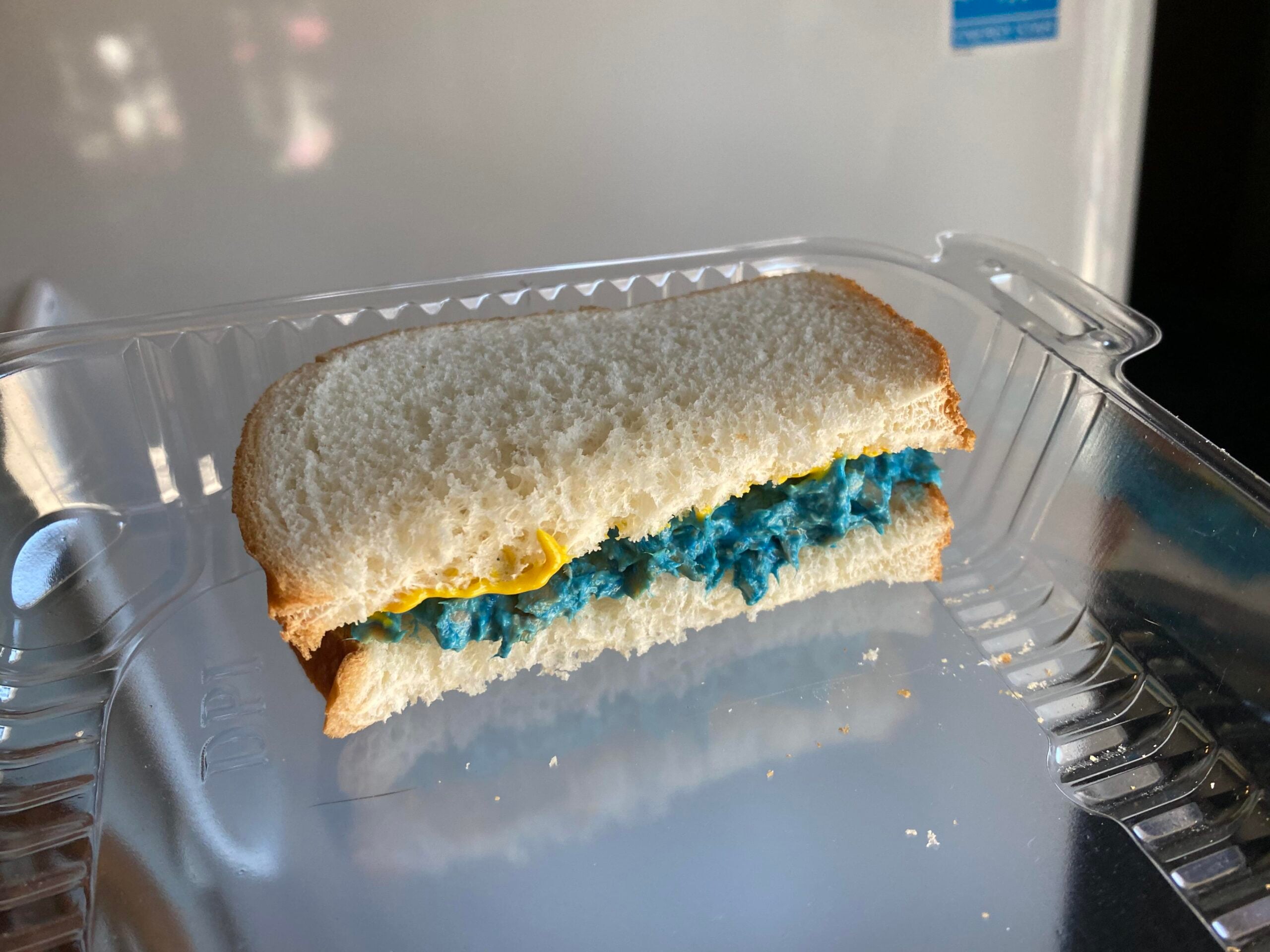 A sandwich on white bread with a blue filling and yellow mustard in a clear plastic container.