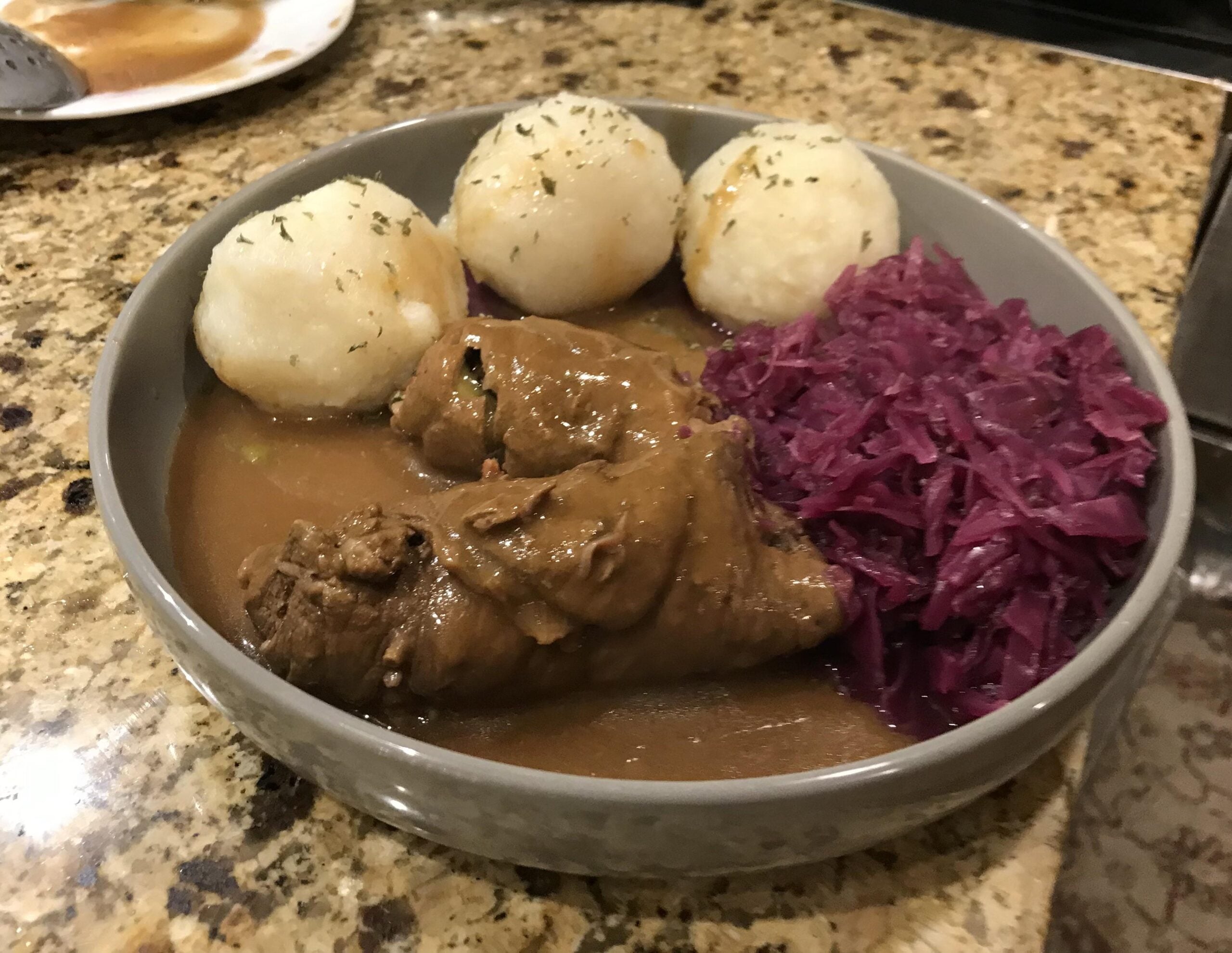 A bowl containing two beef rouladen in gravy, three potato dumplings, and a side of red cabbage.