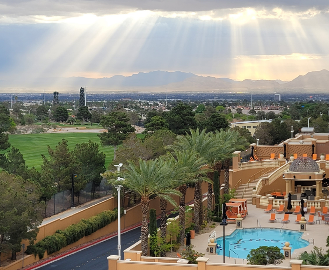 Sunlight streams through clouds over a desert valley, with a golf course, palm trees, and a swimming pool in the foreground.