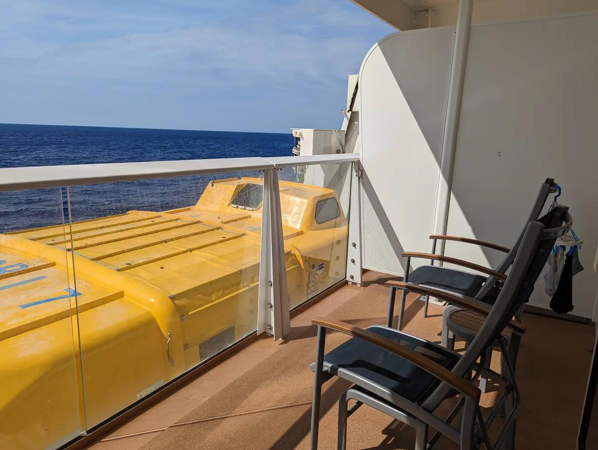 View from a cruise ship balcony featuring two chairs and a large yellow lifeboat outside the glass railing.