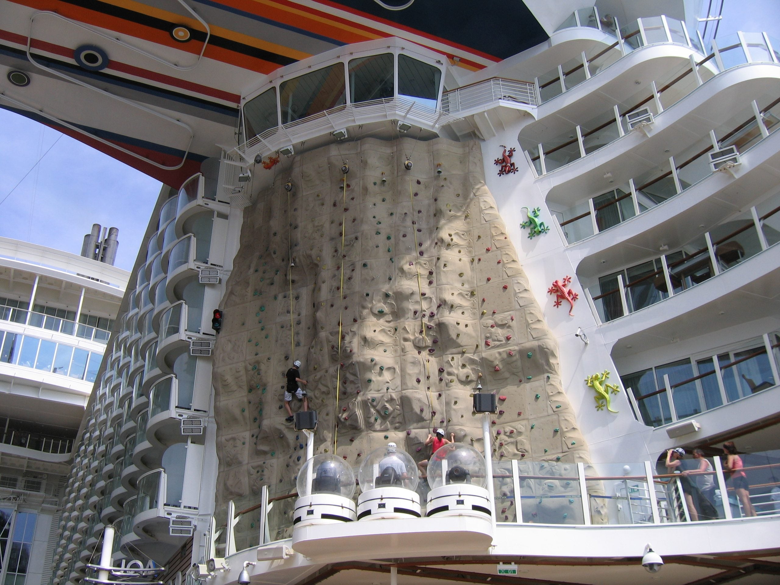 A large outdoor rock climbing wall on a cruise ship, with several people climbing and others watching from the deck. The wall has different colored climbing holds and is surrounded by balconies and glass observation pods.