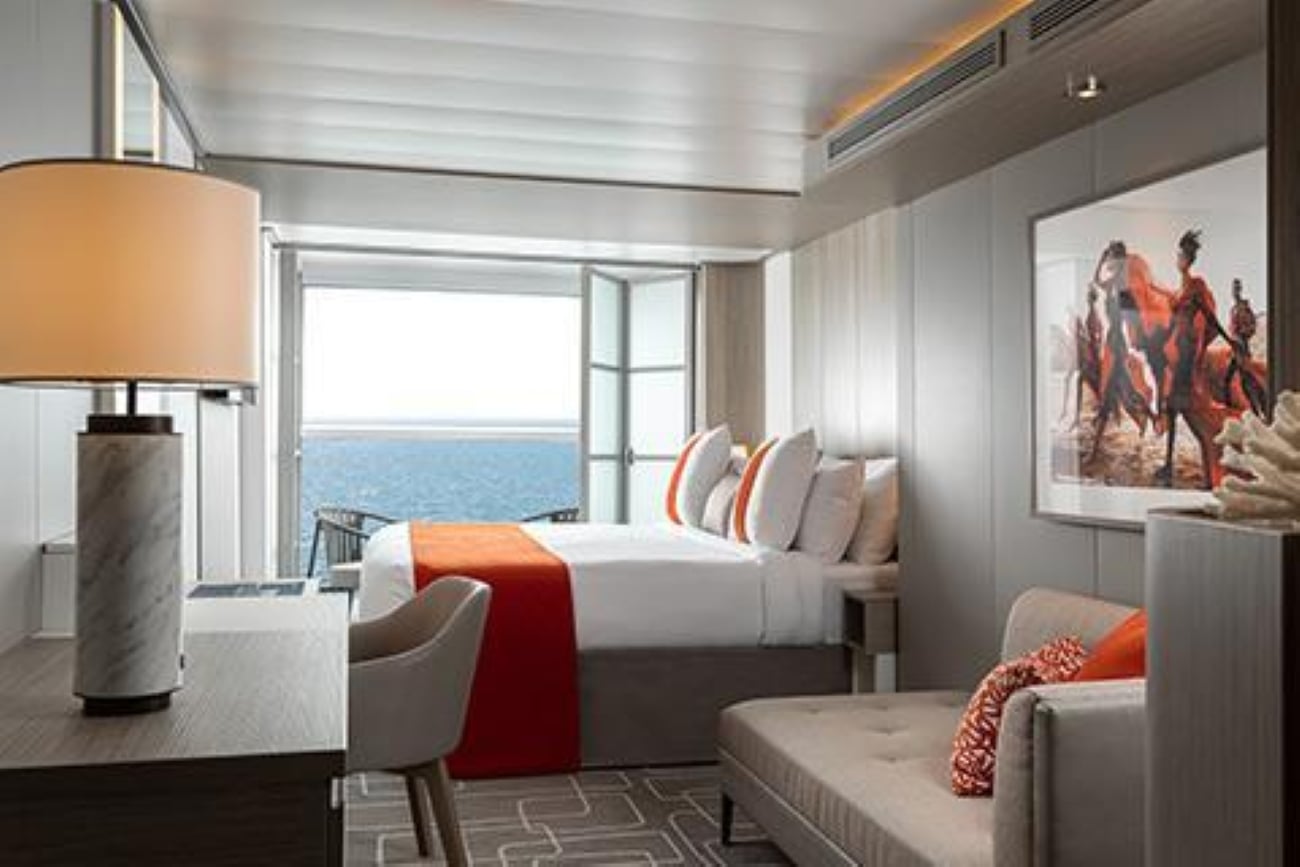 A modern cruise ship cabin featuring a bed with a bright orange runner facing a balcony door with an ocean view, a desk with a lamp, and a lounge chair.