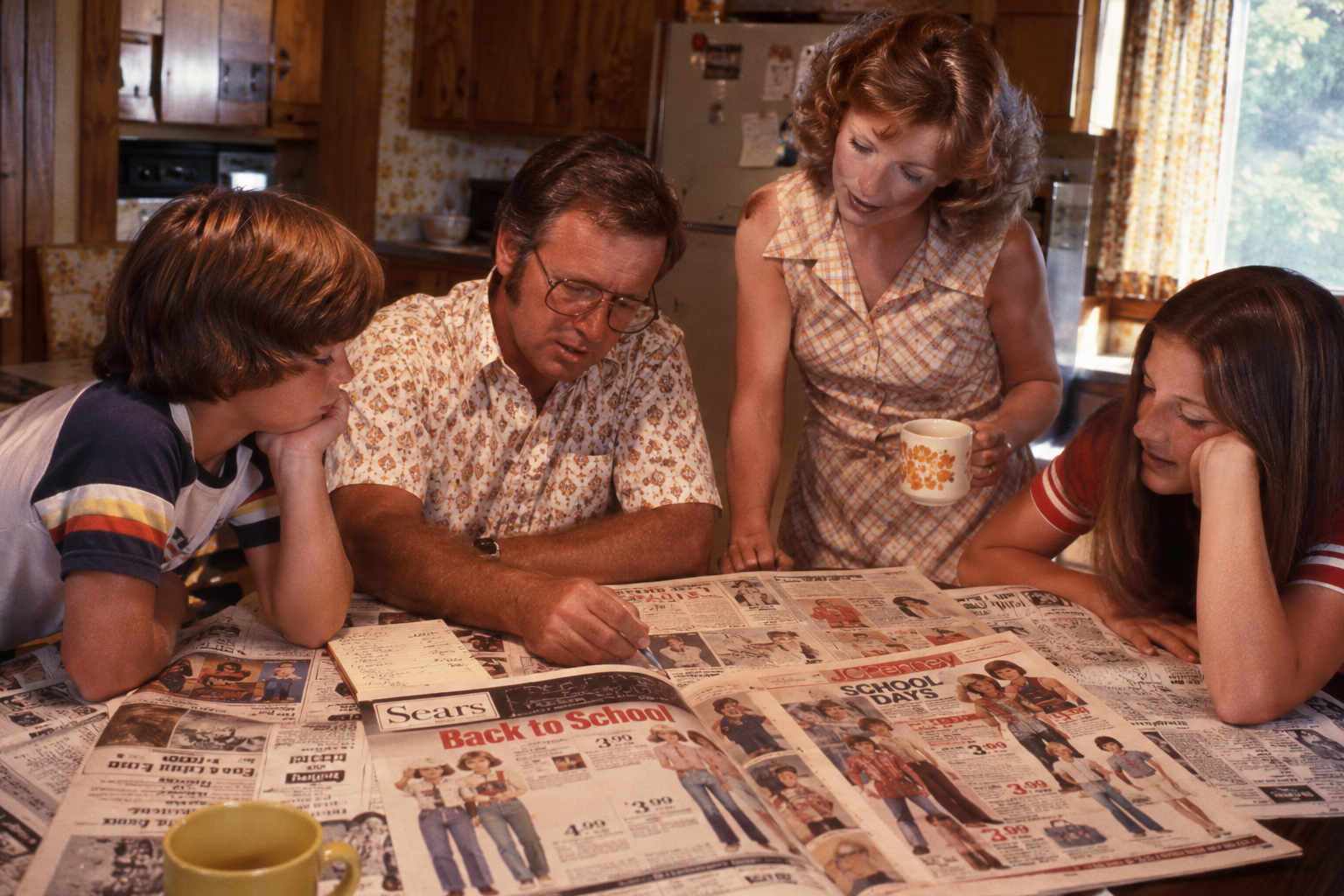 A family of four sits at a kitchen table looking at a Sears back-to-school newspaper advertisement.