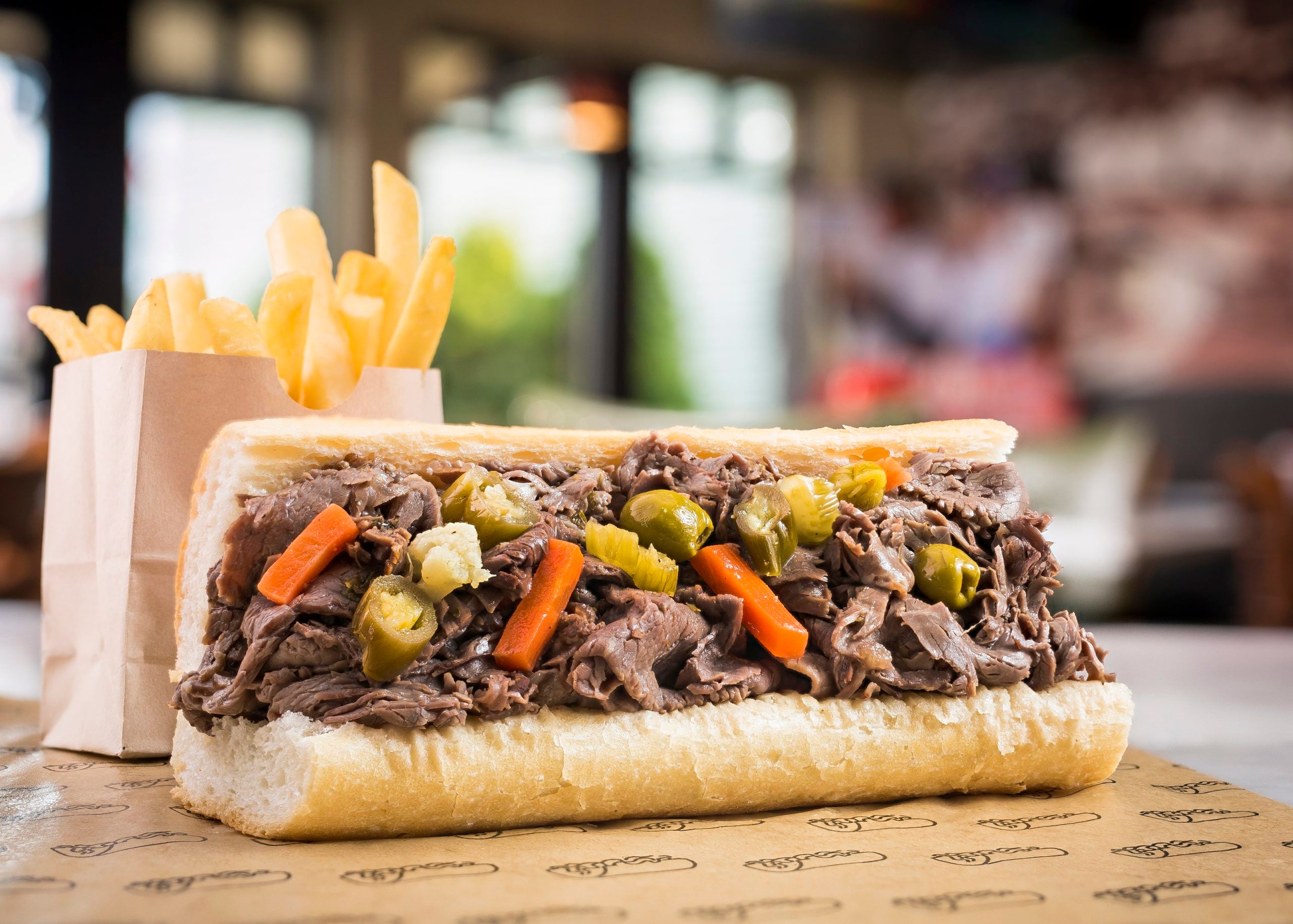 A sandwich filled with sliced beef and pickled vegetables sits on a wrapper next to a paper bag of French fries. The background is blurred, highlighting the food in the foreground.